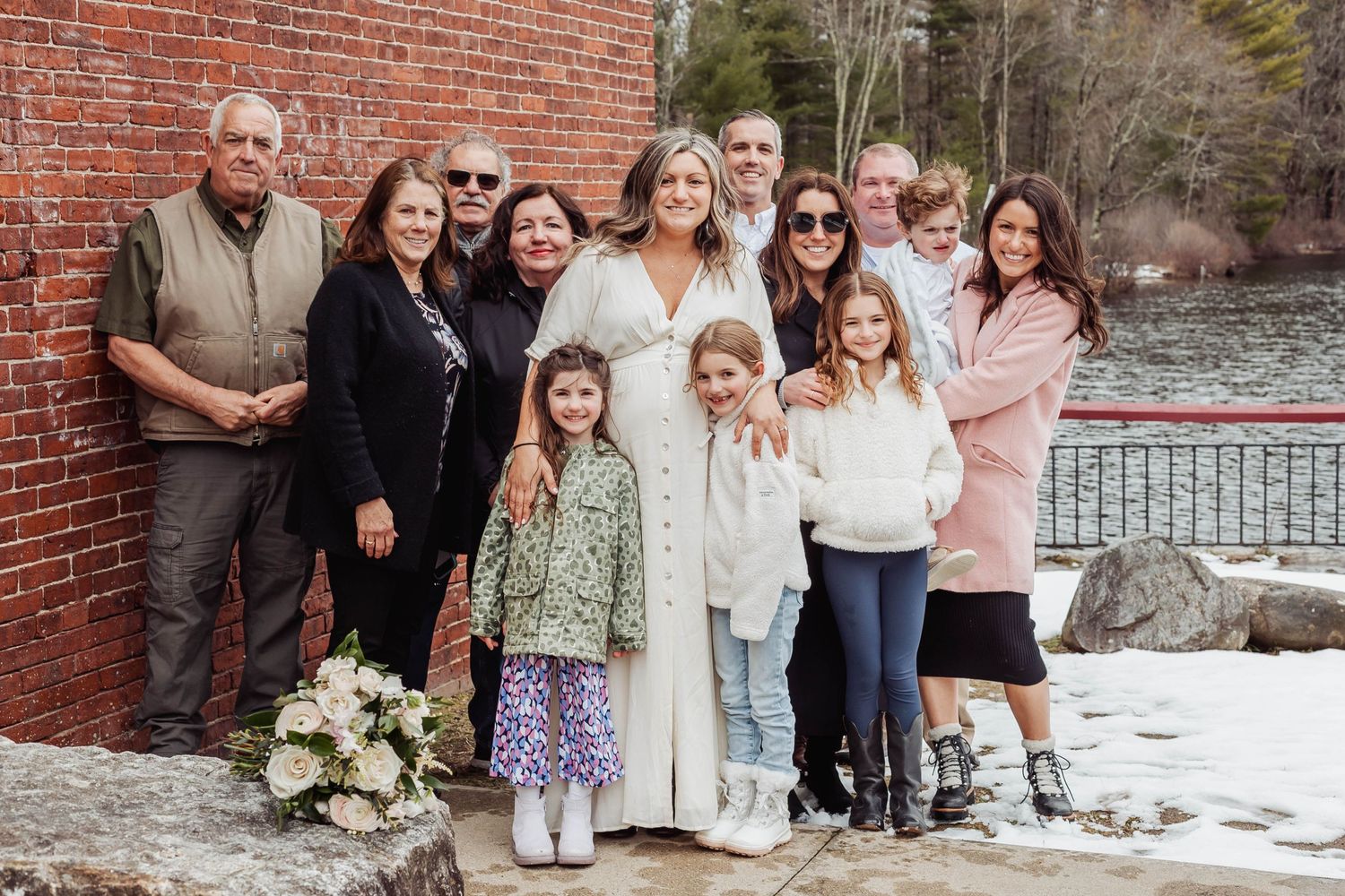 Extended family gathers for a group photo on a snowy deck by a brick building and lake.