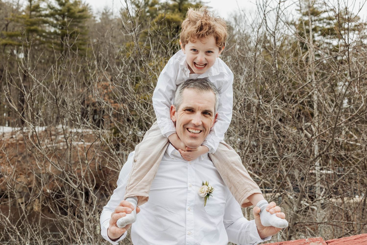 A playful winter moment between two people by a brick building with bare trees in background.