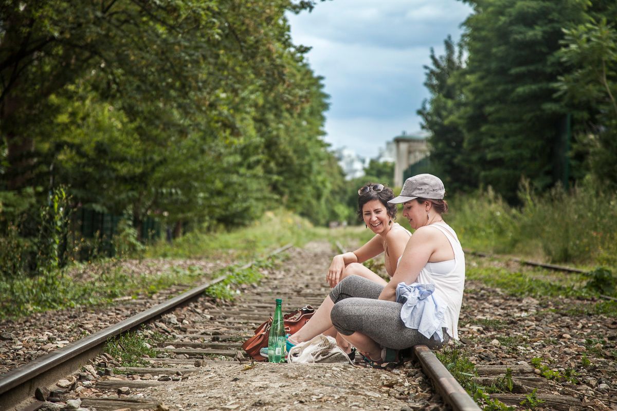 Two figures sit casually on abandoned railroad tracks surrounded by summer foliage.