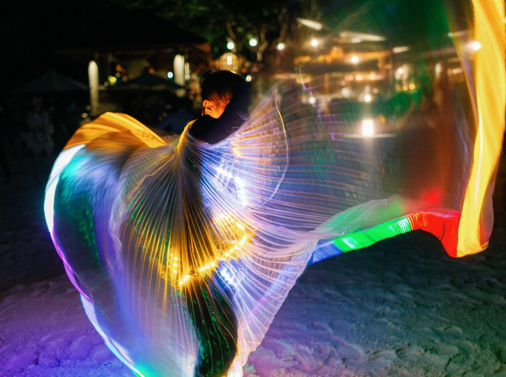 Colorfully dressed man dances with illuminated wings at Amanpulo wedding celebration.