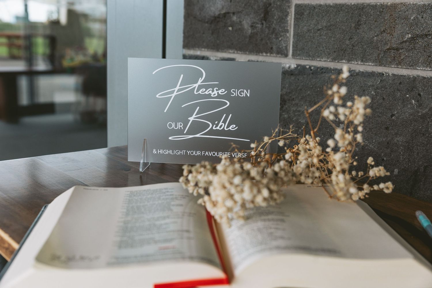 Open Bible with decorative baby's breath flowers and a hand-lettered 'Please sign' note card in soft lighting.