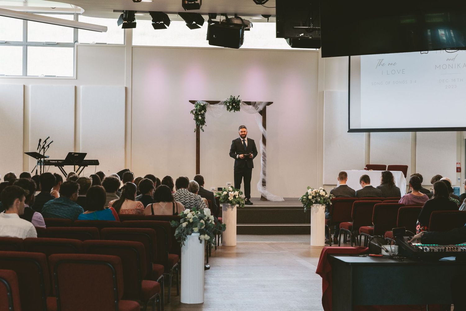 Modern church ceremony setup with white floral arch and rows of wooden pews filled with guests.