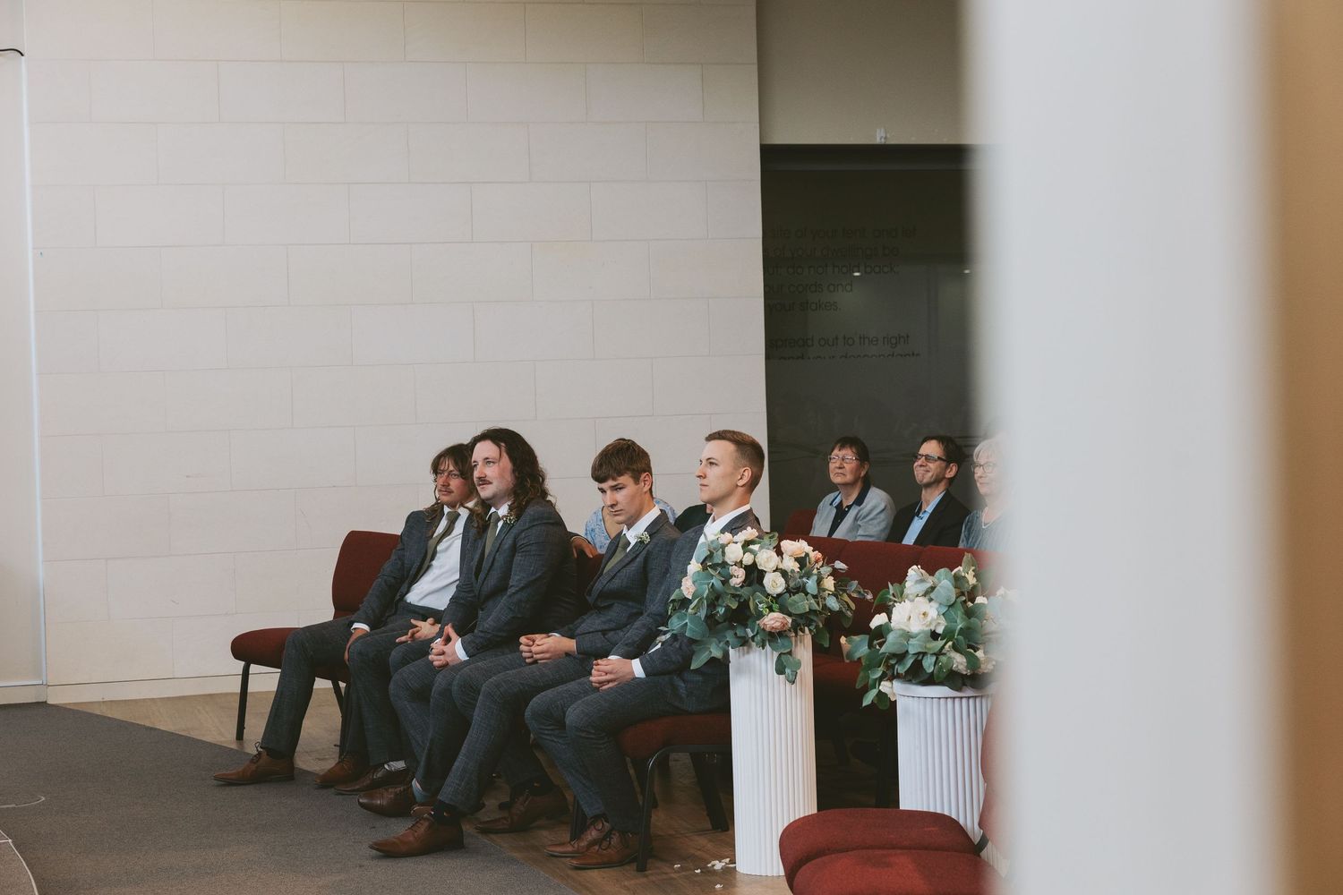 Wedding party members seated on chairs in a minimalist venue space with white floral arrangements.