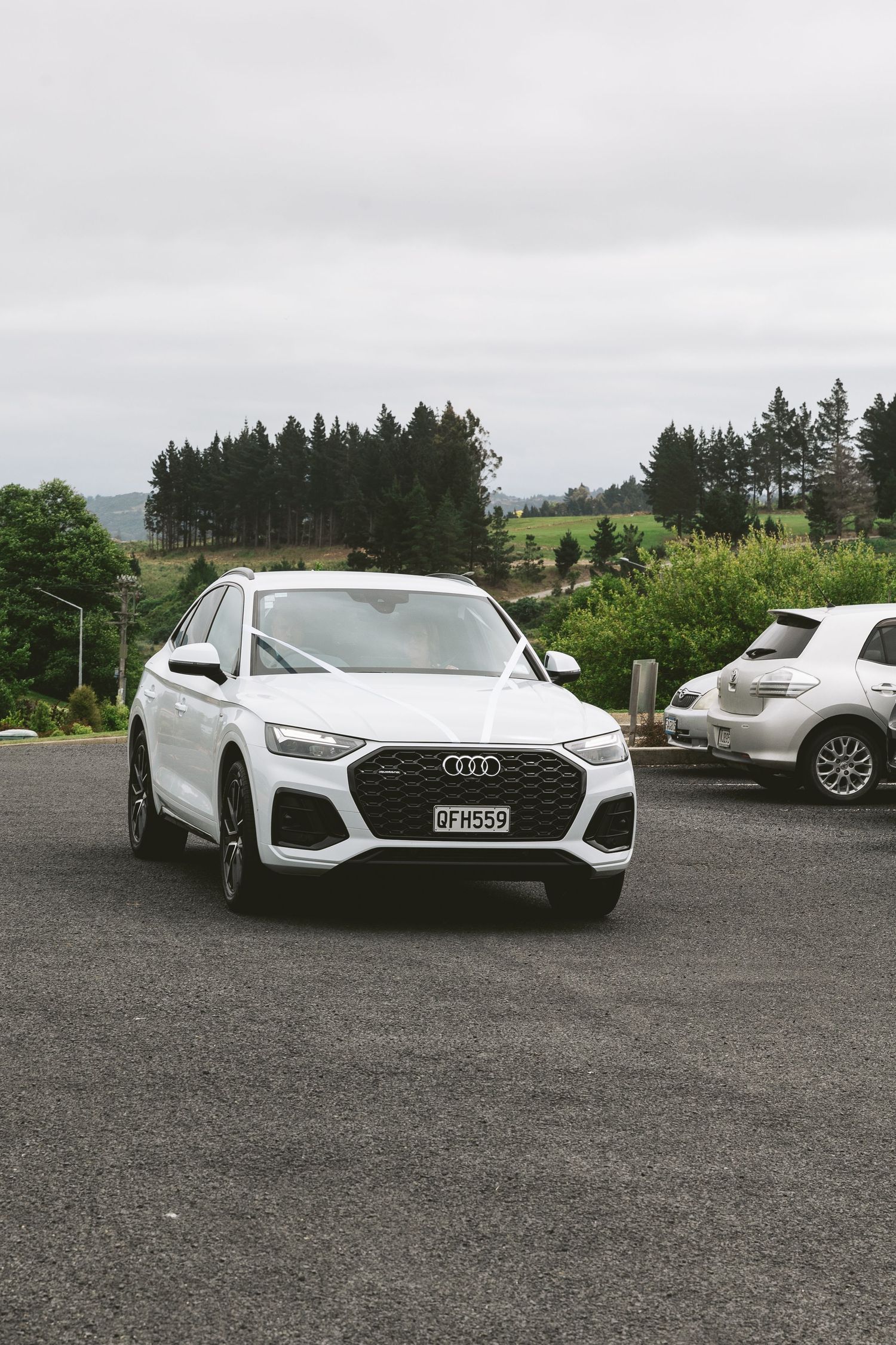 White Audi SUV parked in lot with pine trees and rolling hills in background on overcast day.