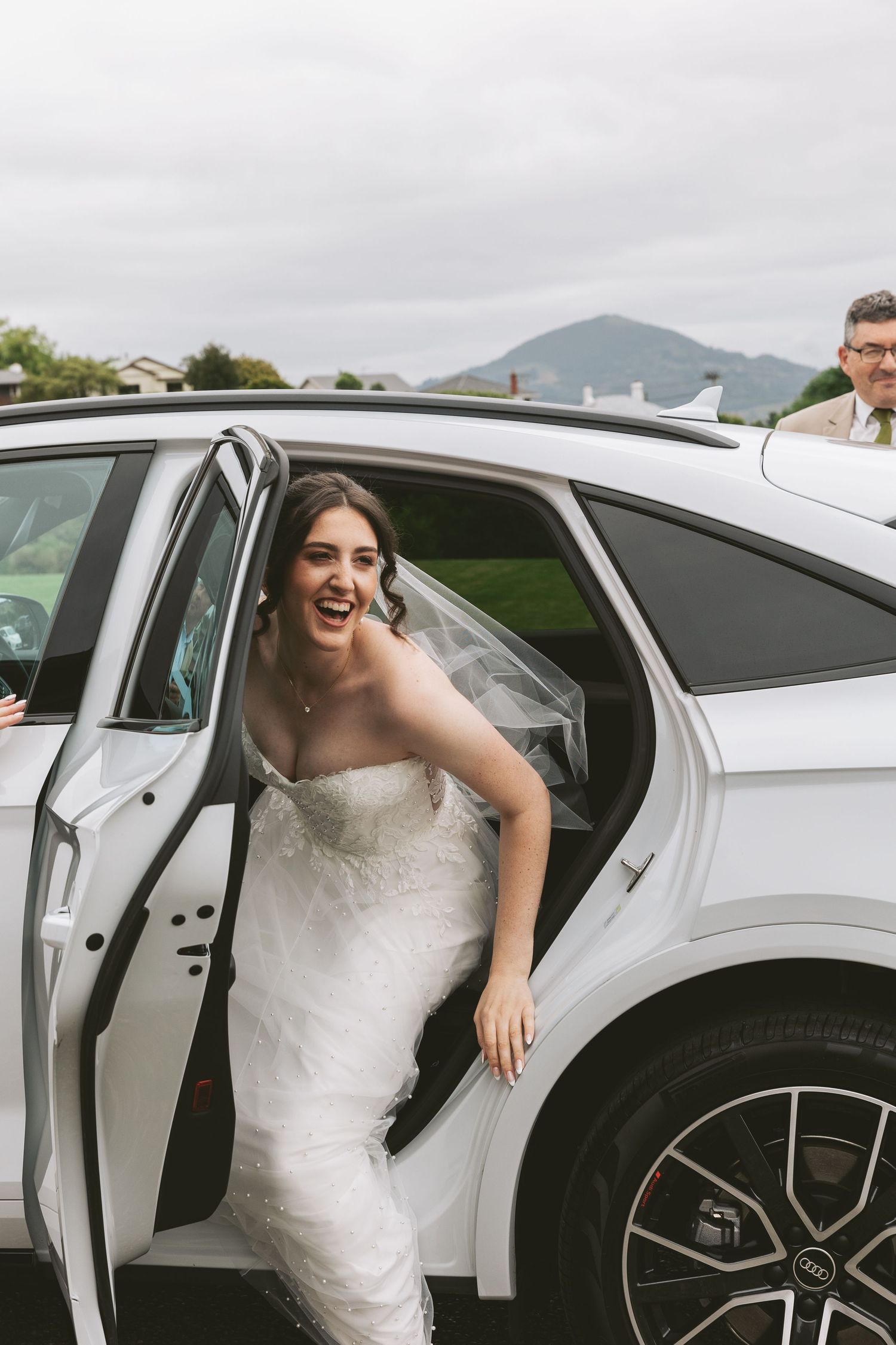Wedding attire and luxury white vehicle against mountain backdrop on cloudy day.