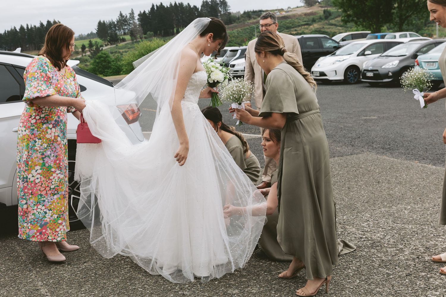 Wedding party helps adjust a flowing white dress in parking lot with mountains visible.