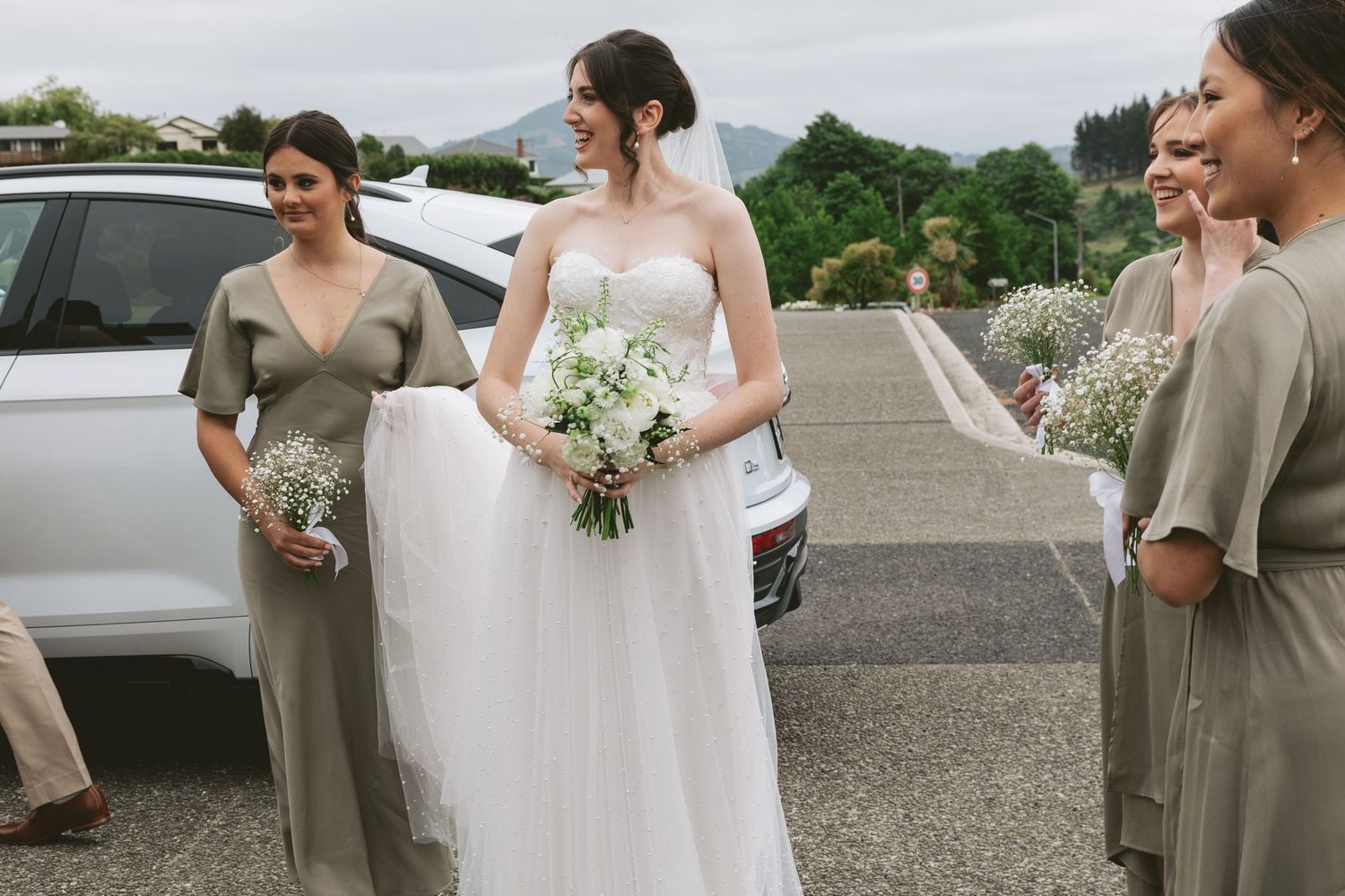 Wedding party in sage green dresses holding white bouquets outdoors.