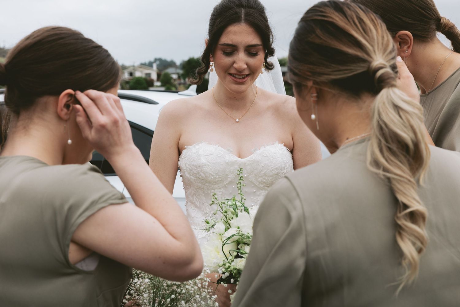 Members of the wedding party in taupe dresses gather around a person in a white gown with delicate floral bouquet.