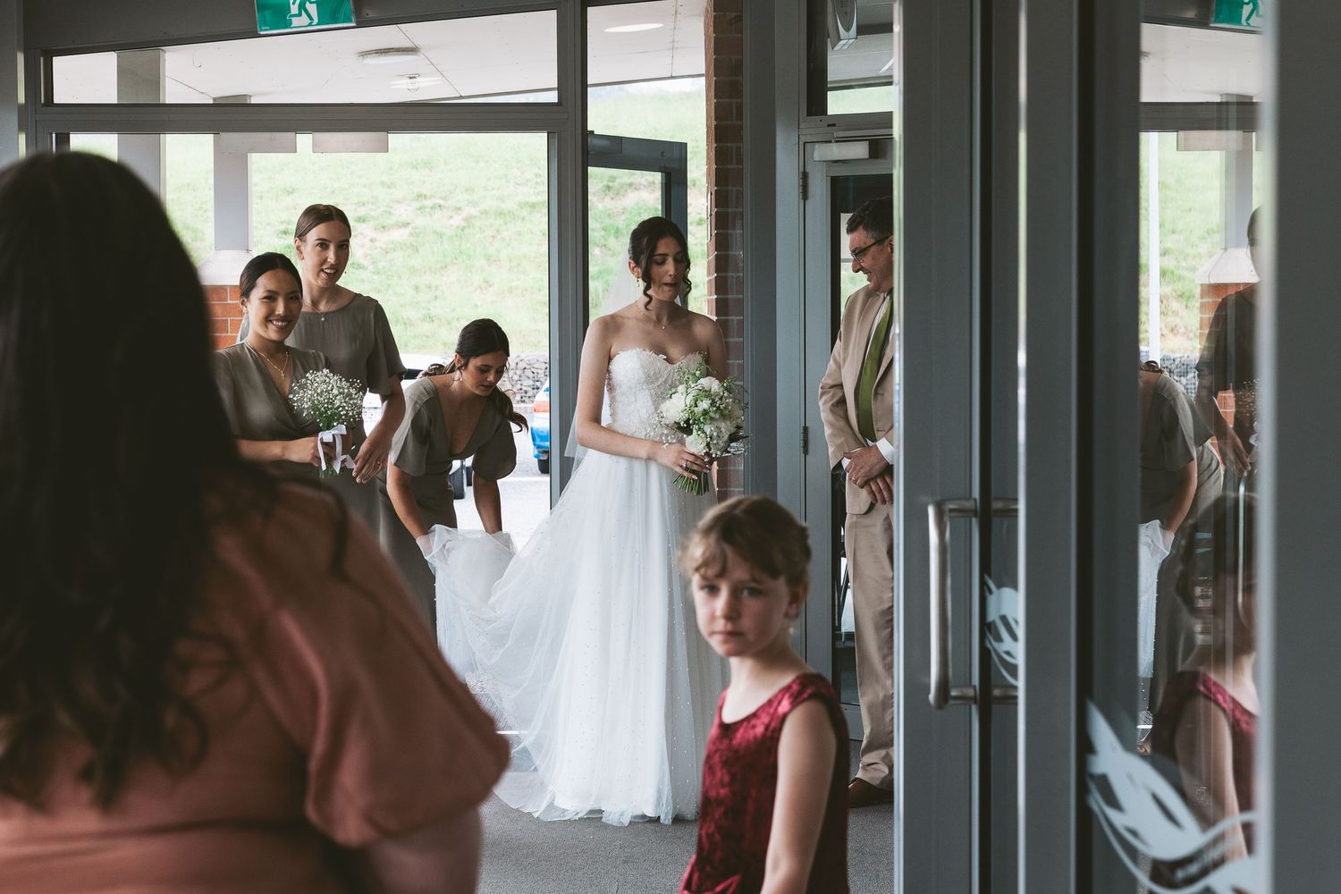 Wedding party members enter a modern building lobby through glass doors.