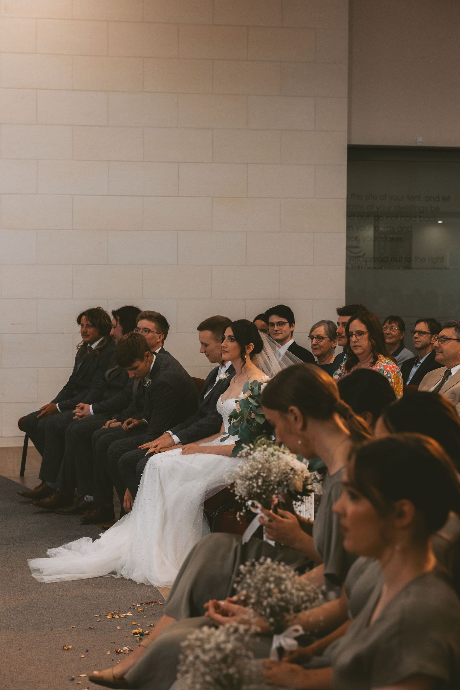 Wedding party members seated in formal attire holding baby's breath bouquets against white brick wall.