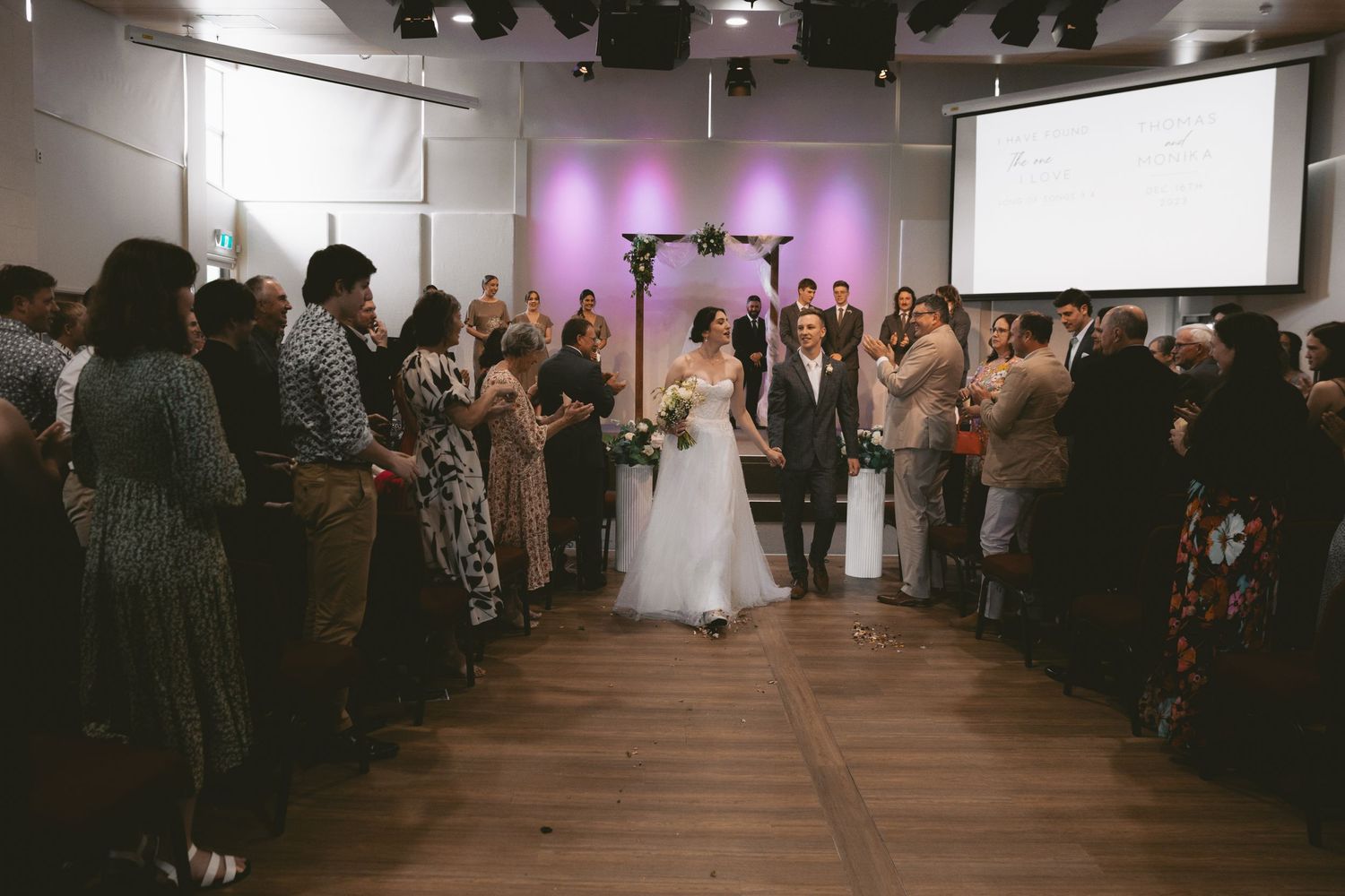Wedding celebration in modern venue with purple accent lighting and guests lining the aisle.