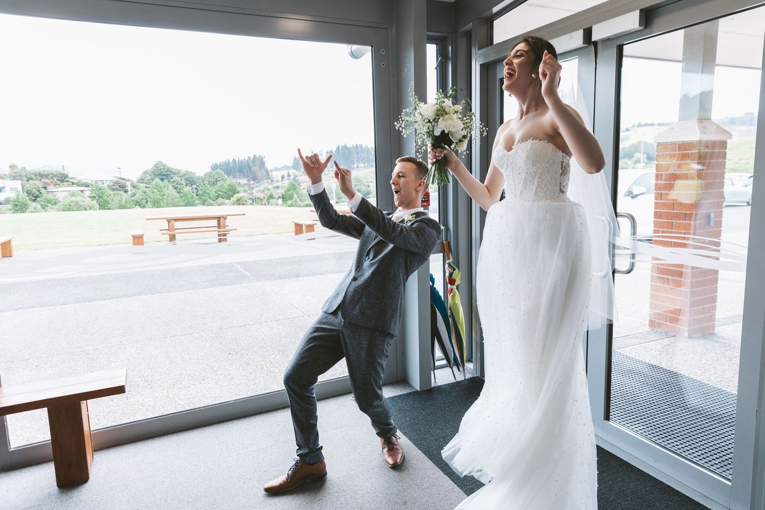 Playful wedding photo of couple posing energetically by glass windows.