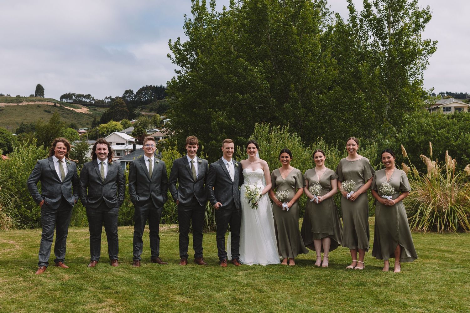 Wedding party poses on grassy lawn with bridal party in gray dresses and groomsmen in matching suits.