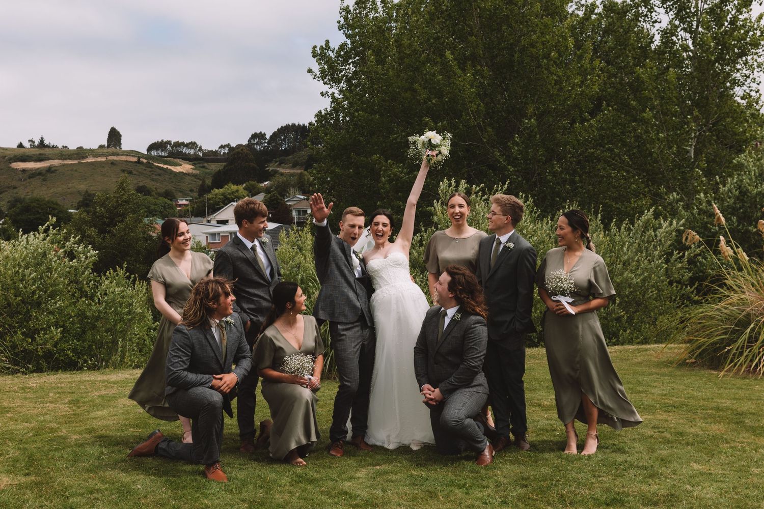 Wedding party strikes fun pose together on grass with trees in background.