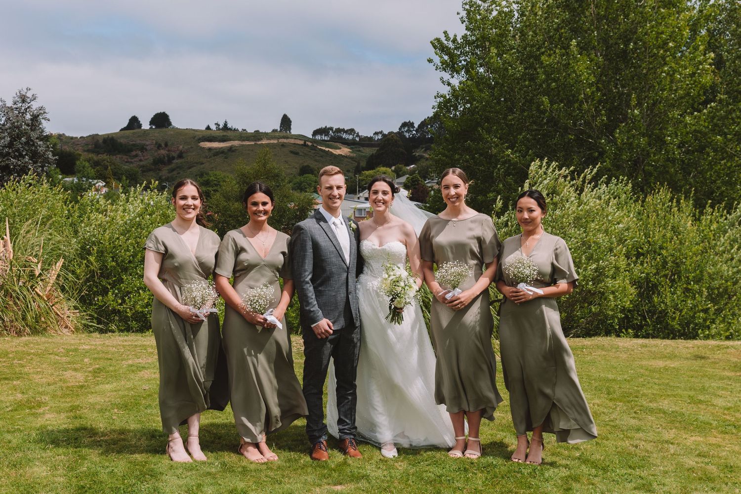 Wedding party group photo with sage green bridesmaid dresses against natural outdoor backdrop.