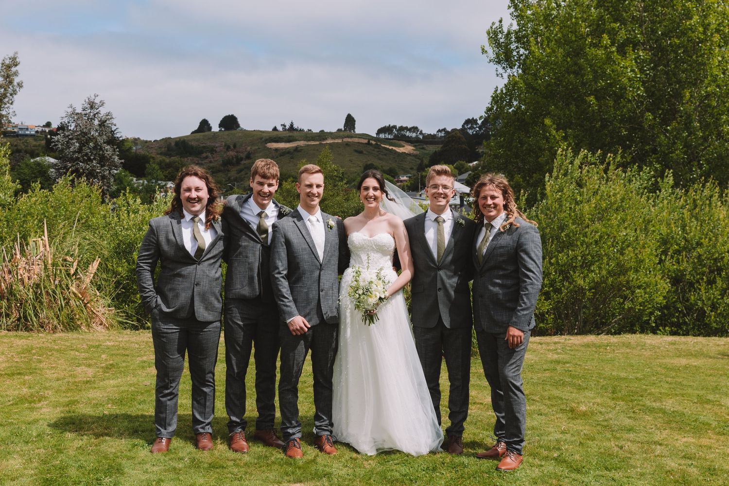 Formal wedding party portrait with matching gray suits and white gown on green lawn.