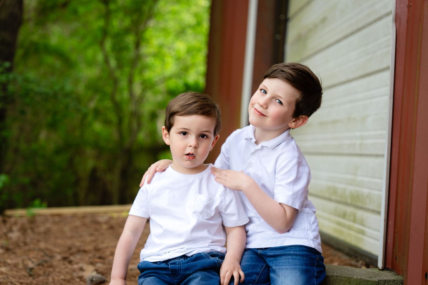 Two young boys in white shirts sitting together outdoors by a white wall.
