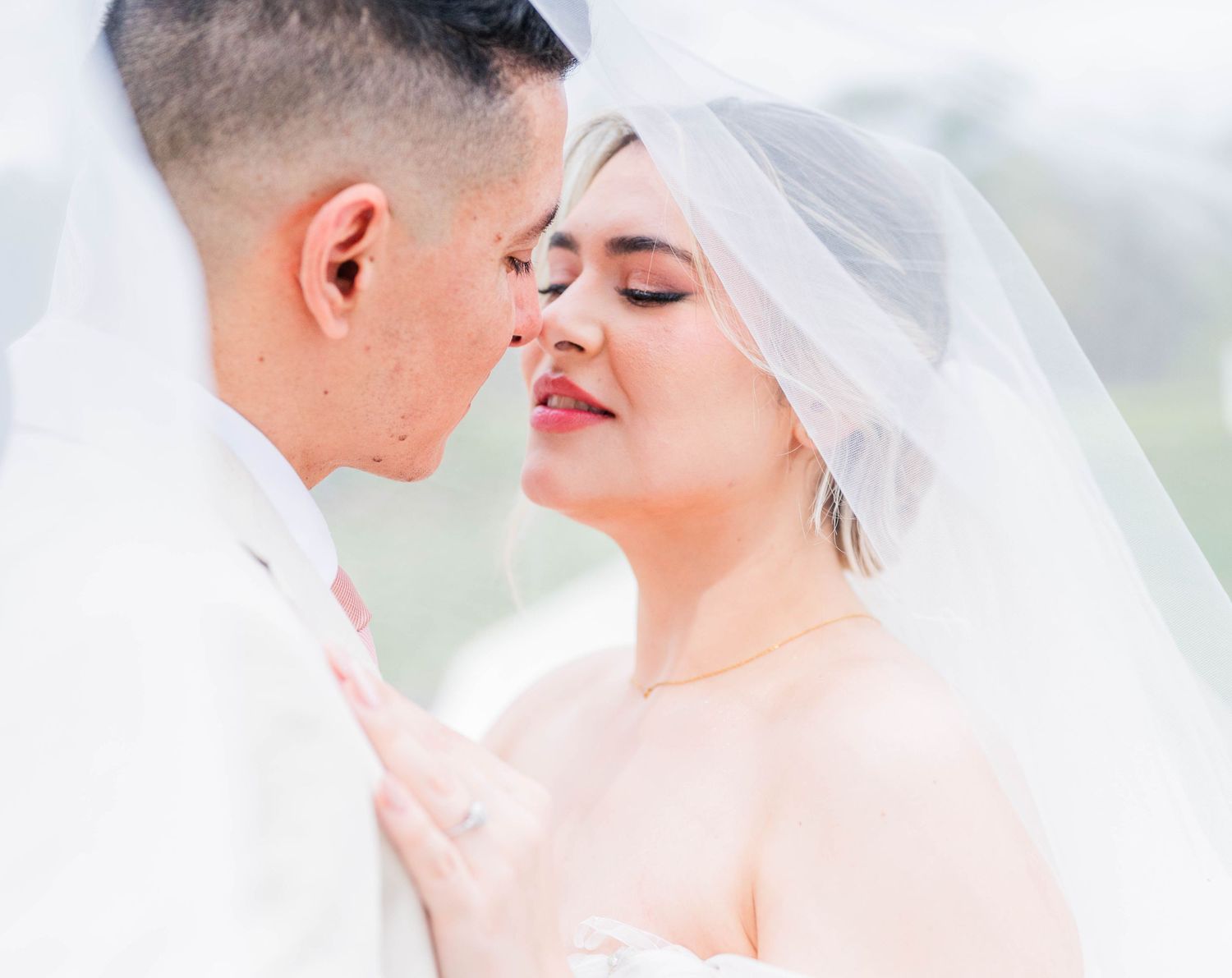 A tender moment between a bride and groom in white, touching foreheads under a soft veil with eyes closed.