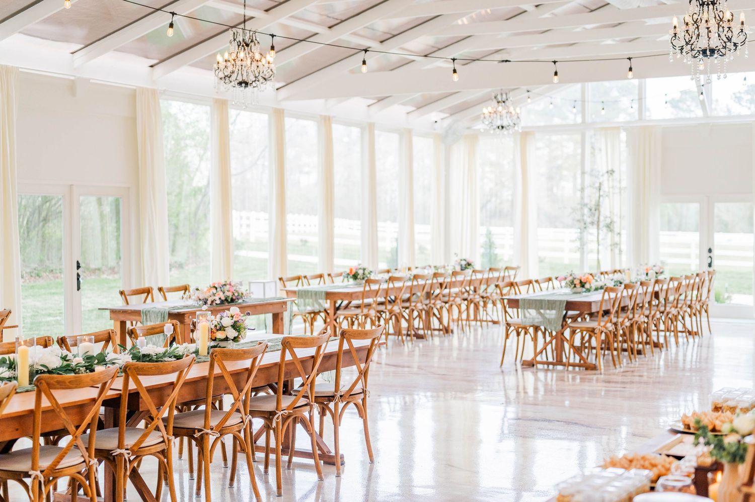 Bright white event venue with wooden cross-back chairs and crystal chandeliers features sunlit windows and floral centerpieces.
