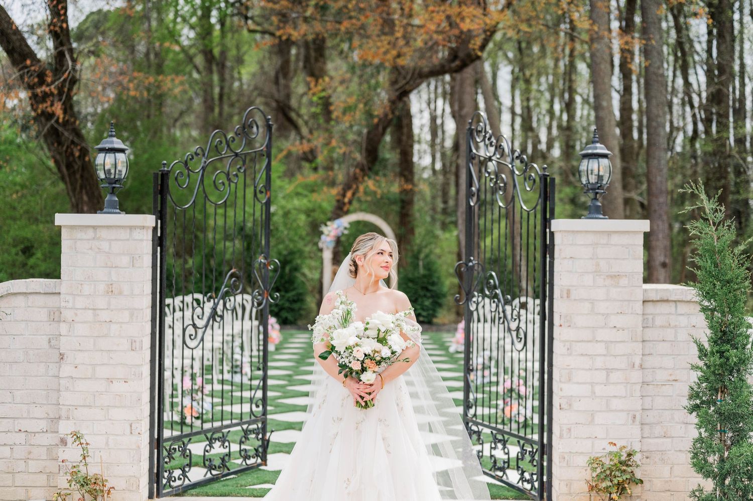An elegant bride in a white dress stands before ornate garden gates with stone pillars and holds a blush floral bouquet.