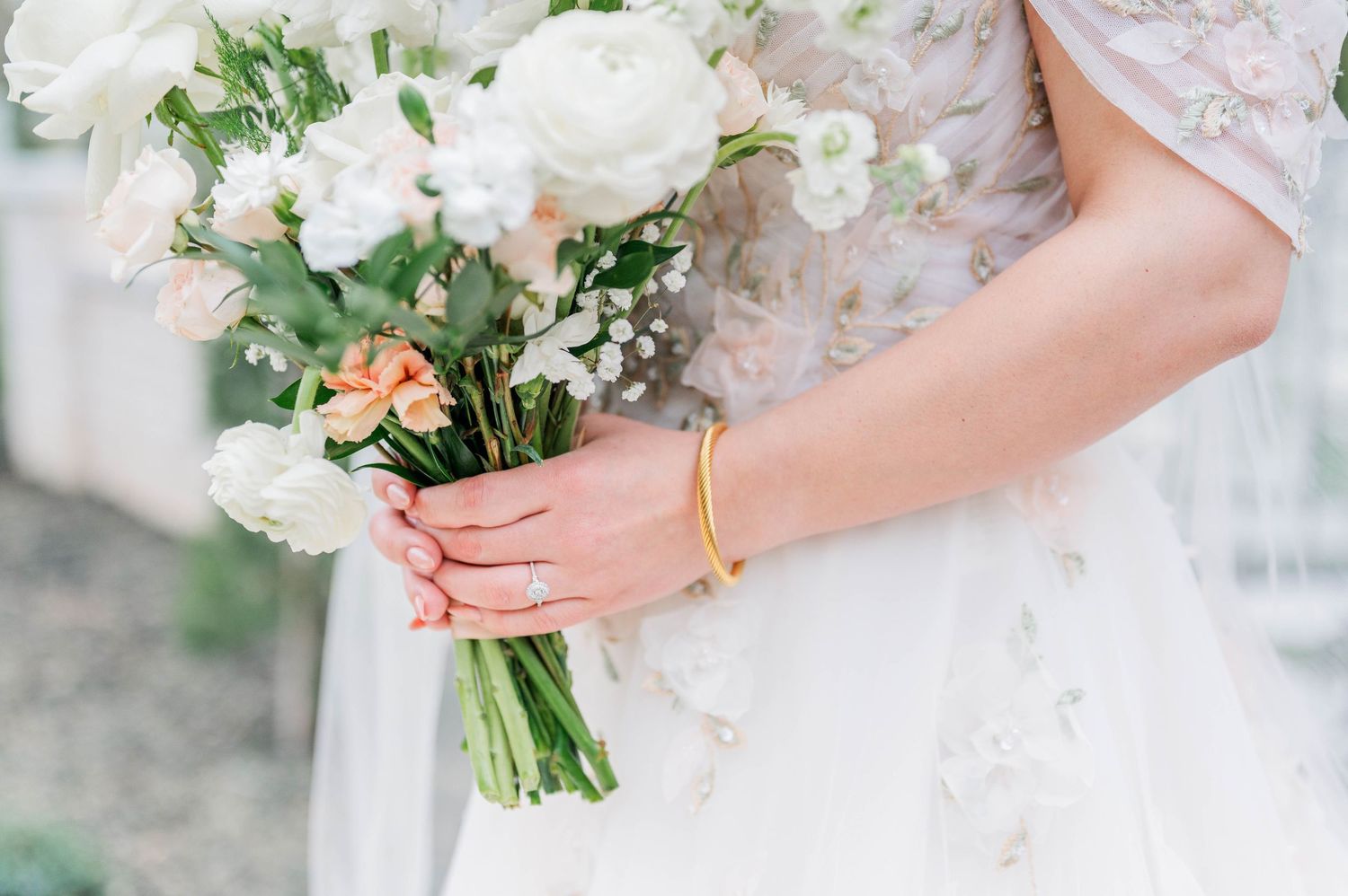 A delicate white and blush pink wedding bouquet held by hands wearing a lace sleeve and gold bracelet.