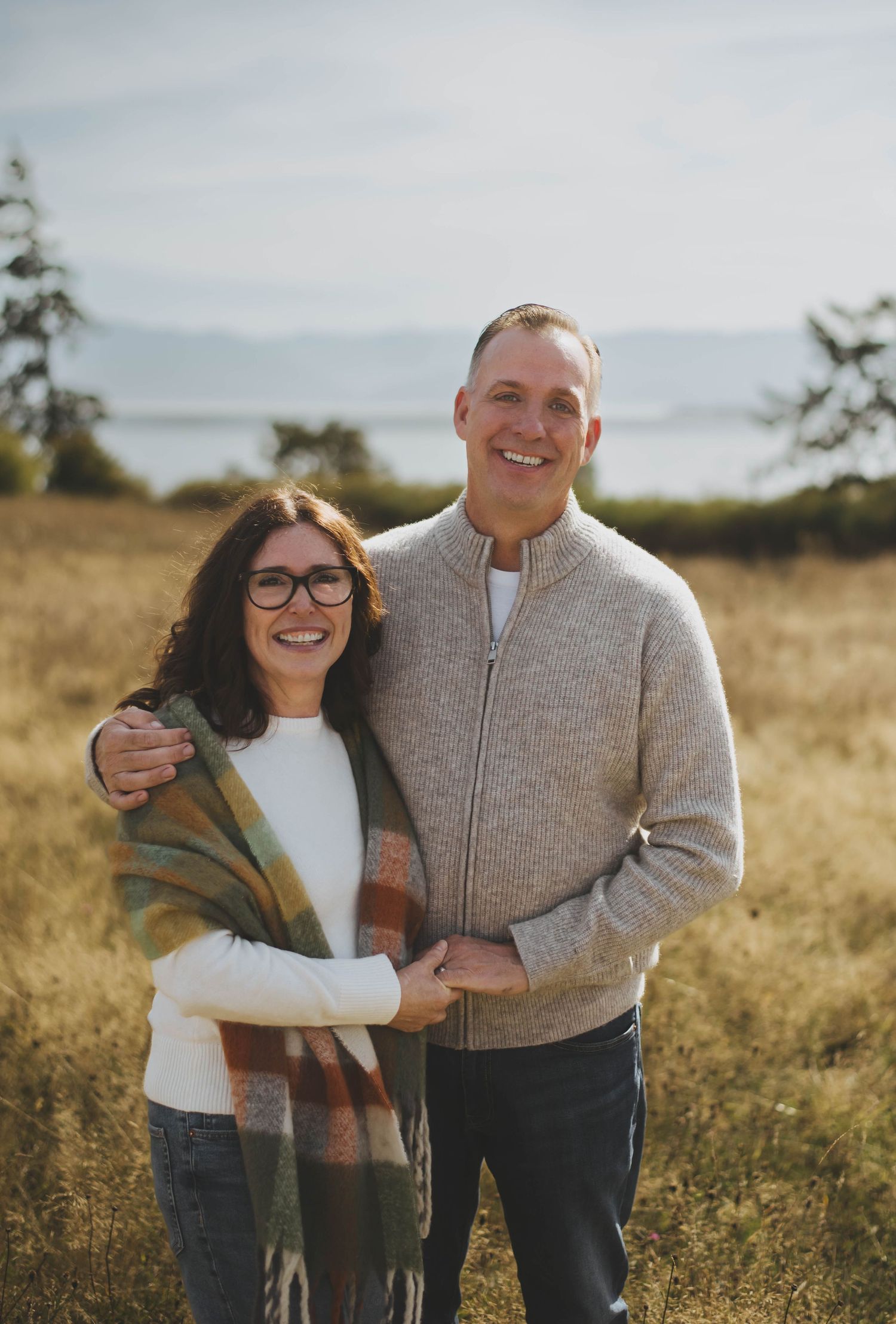 A couple stands closely together, smiling warmly in a sunlit field, wrapped in cozy sweaters with an ocean background.