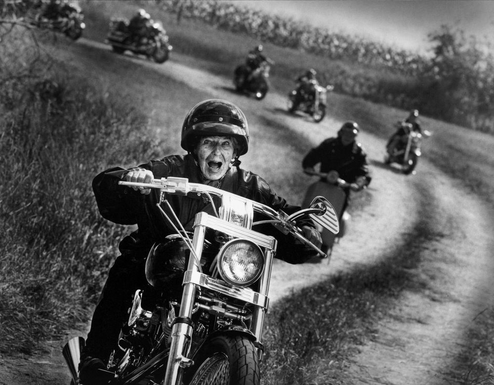 A group of motorcyclists ride along a dusty dirt road in dramatic black and white vintage photography.