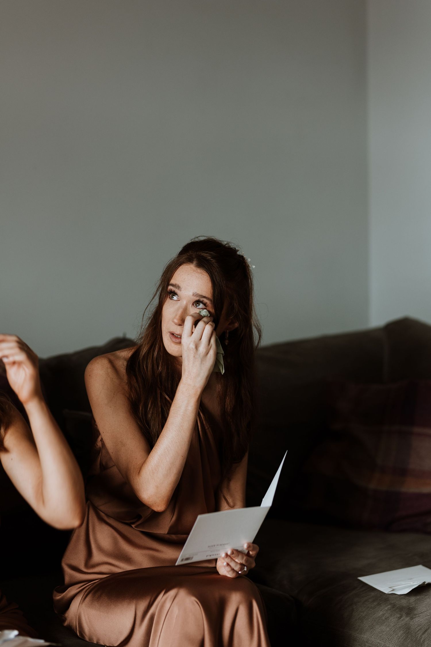 Bridesmaid in a silk dress sits on a dark couch while emotionally reading letter written by bride and groom before ceremony.