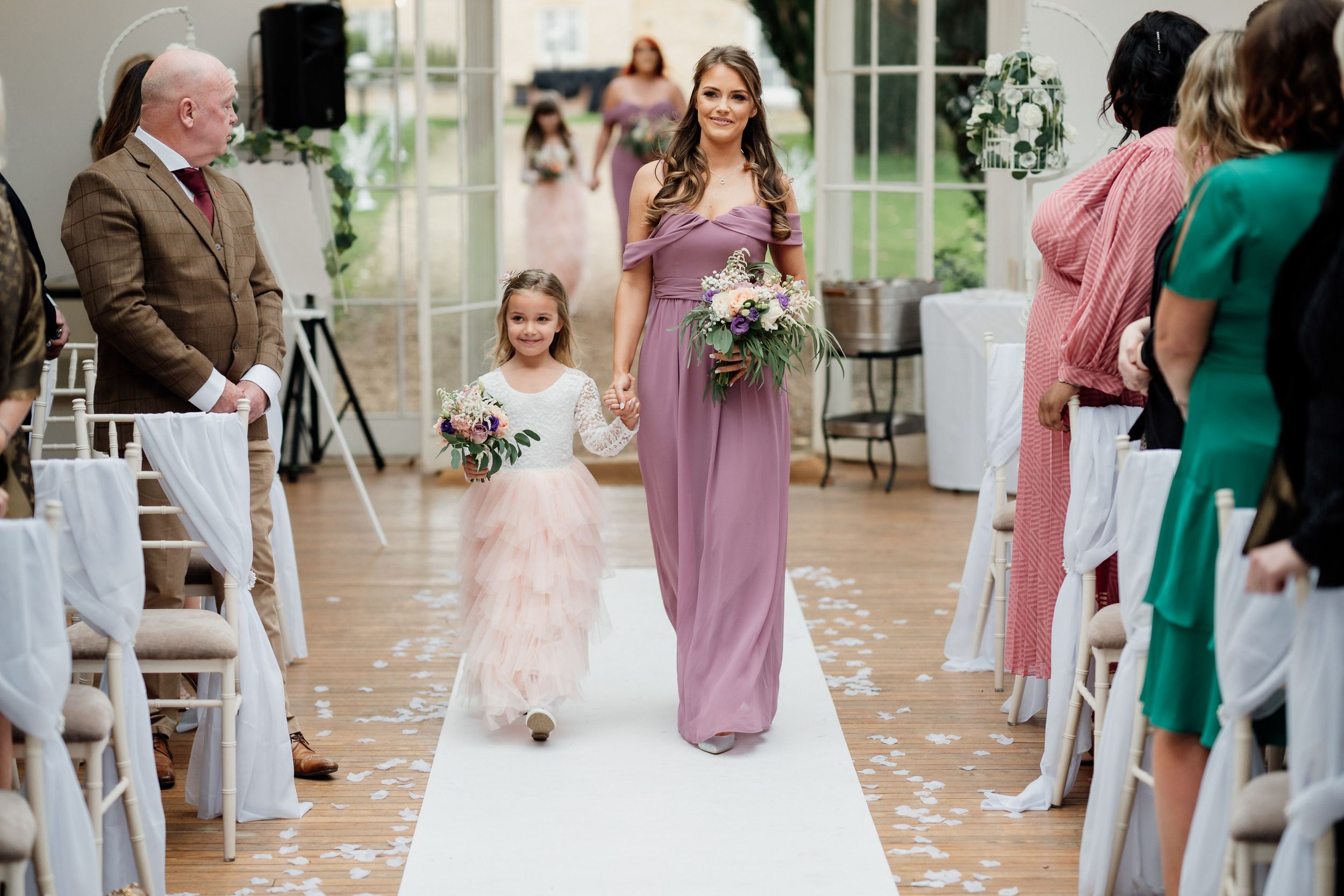 Bridesmaid in mauve dress and flower girl in pink walk down white aisle.