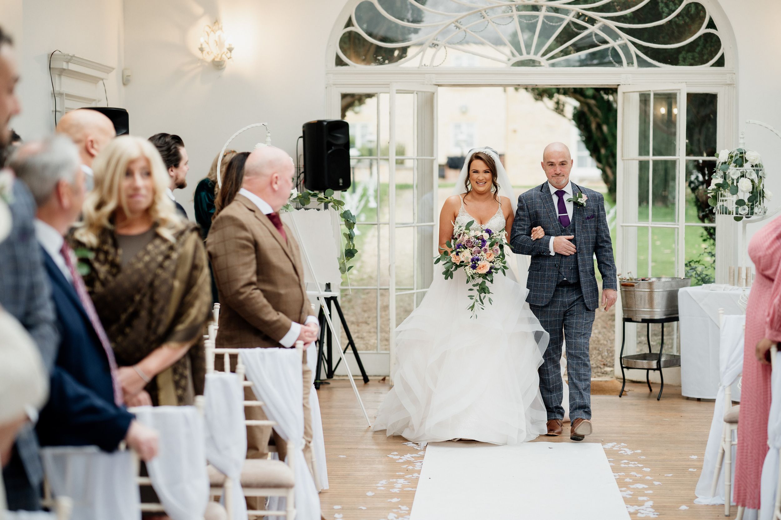 Wedding processional in an elegant white room with arched windows and chandeliers.