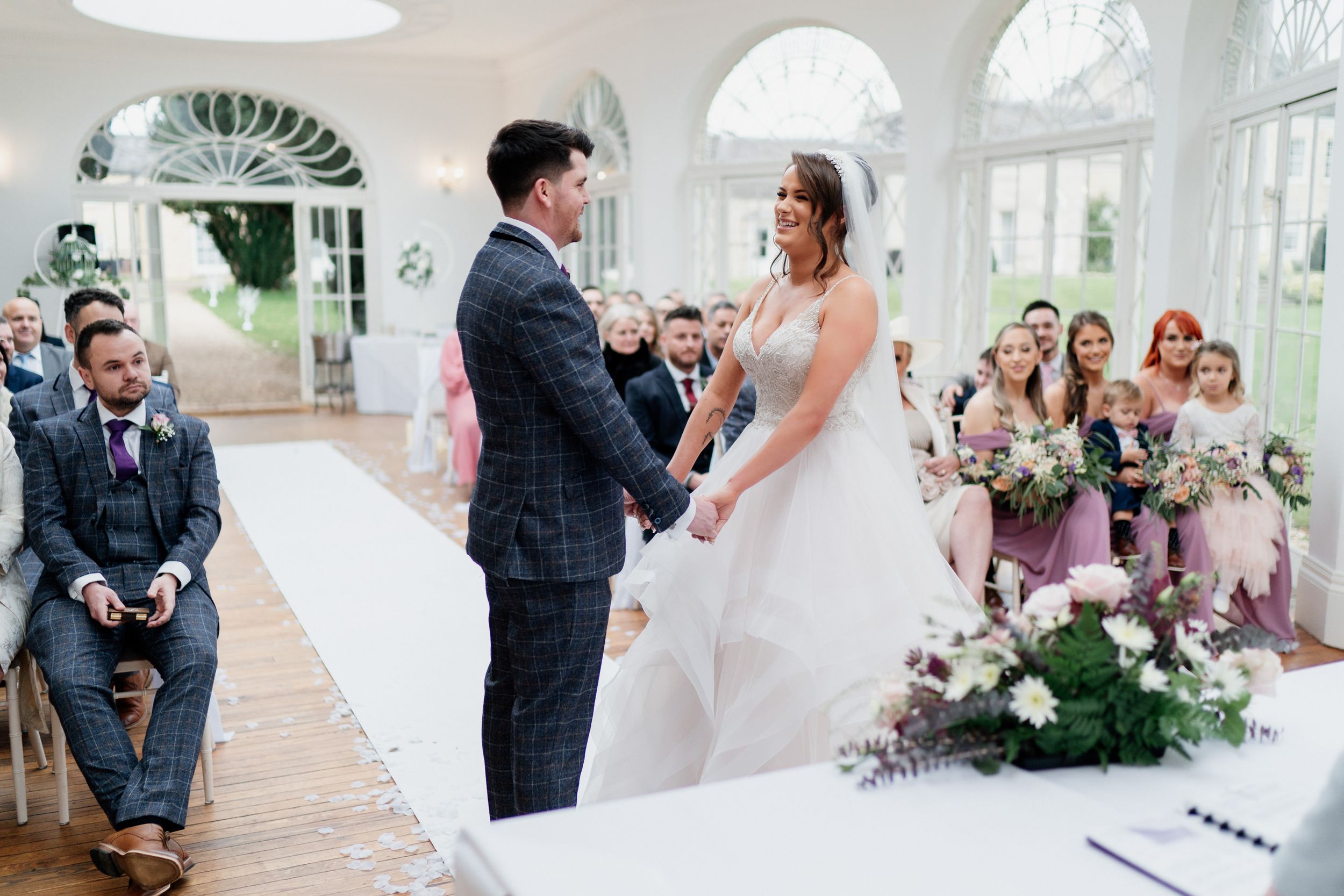 Wedding ceremony with floral decorations in bright indoor venue with arched windows.