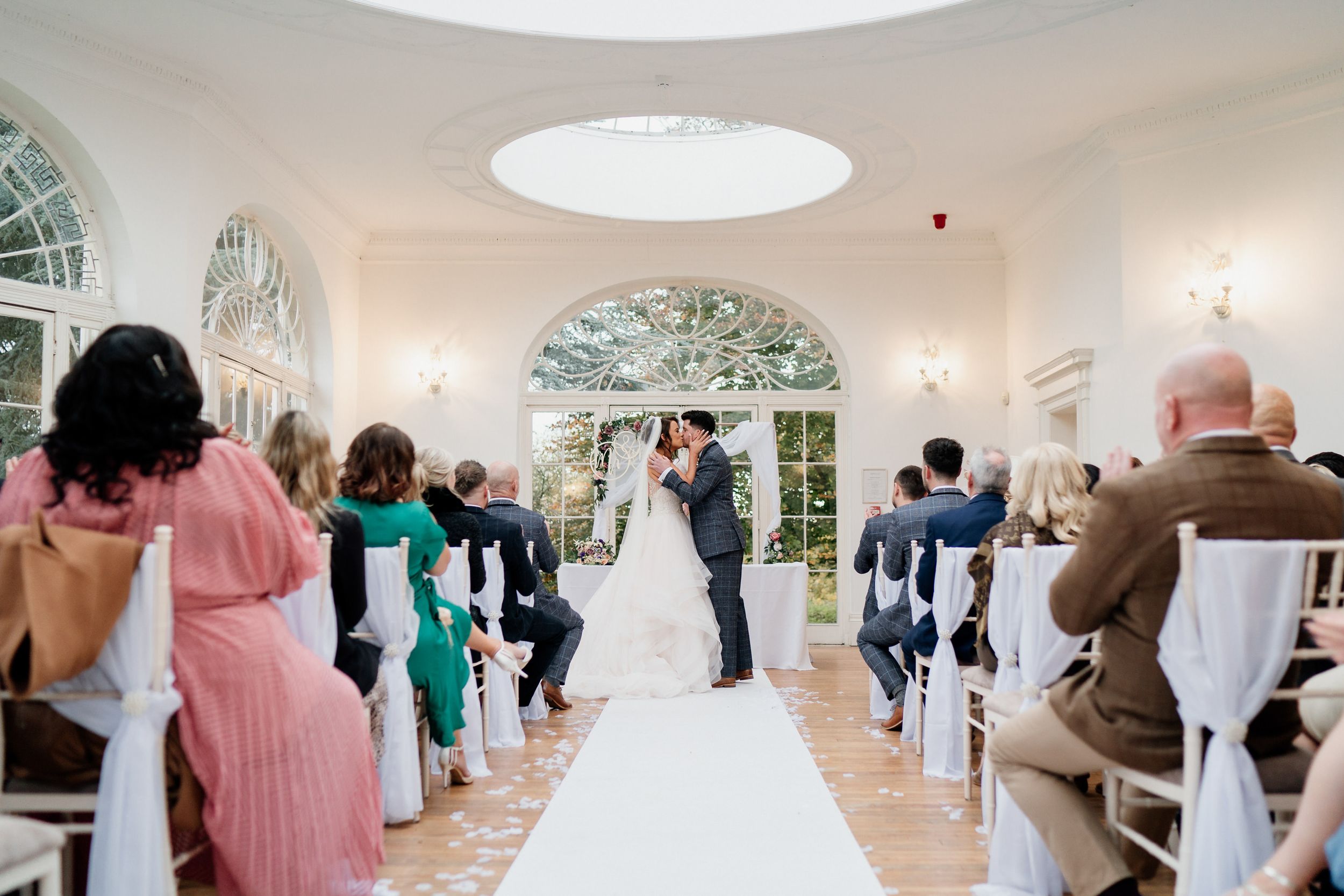 Wedding ceremony at Barton Hall in a bright sunlit room with arched windows and guests seated in white chairs.