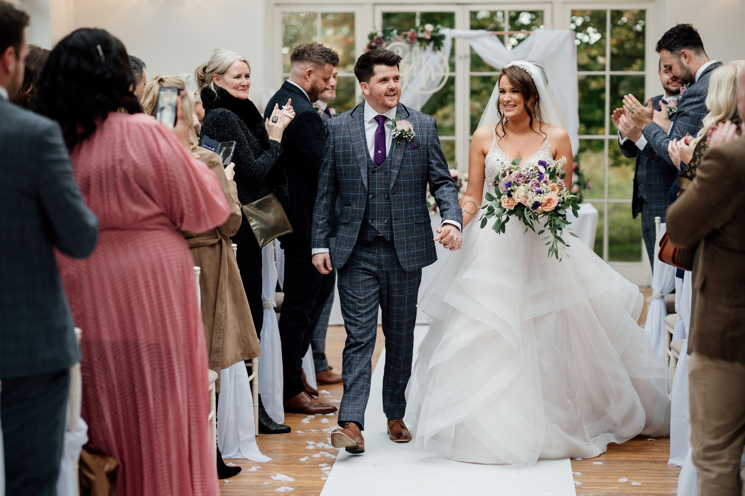 Newlyweds walking down the aisle while guests cheer in a light-filled ceremony space.