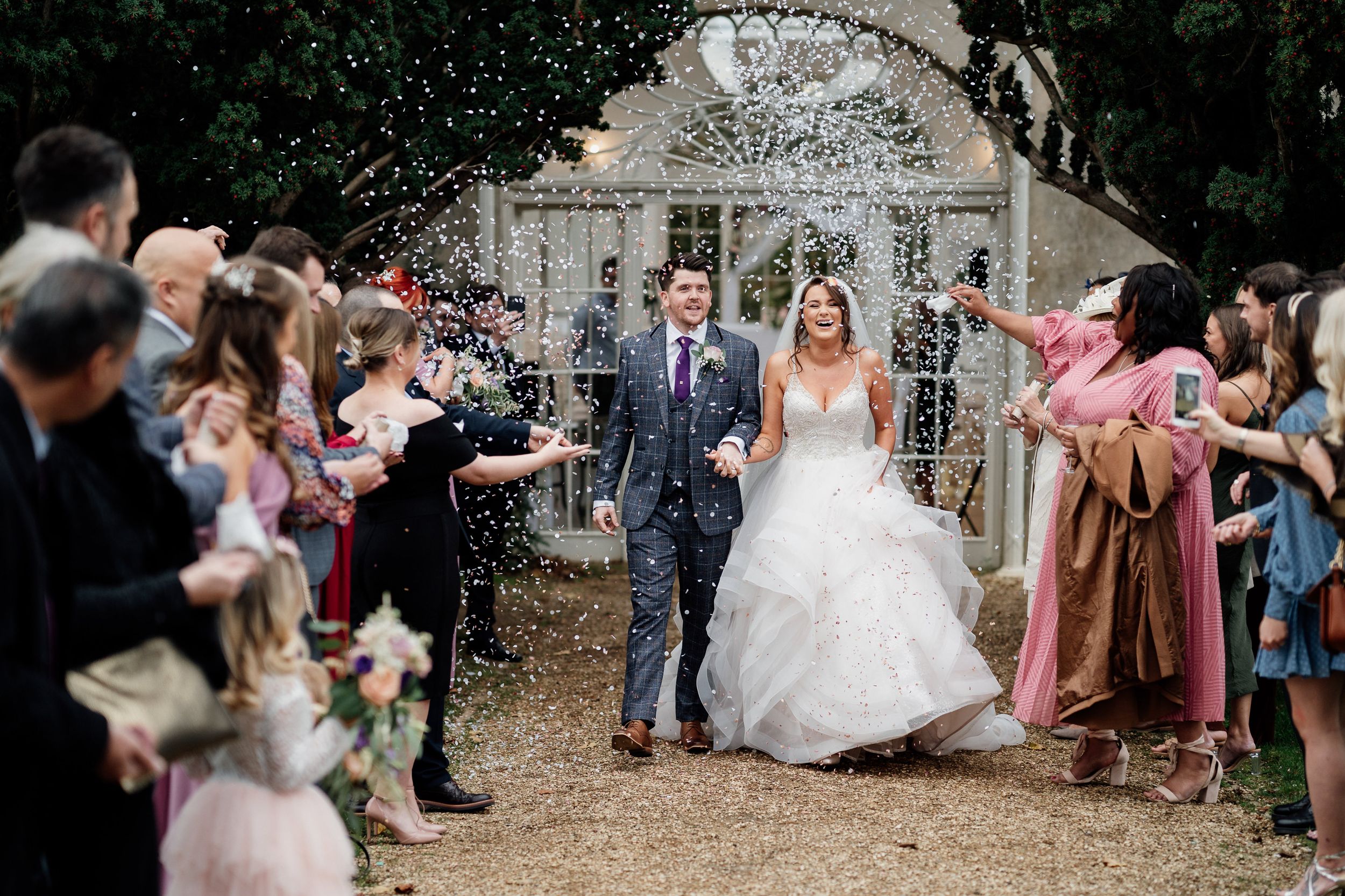 Couple exits through an outdoor archway while guests throw confetti in celebration.