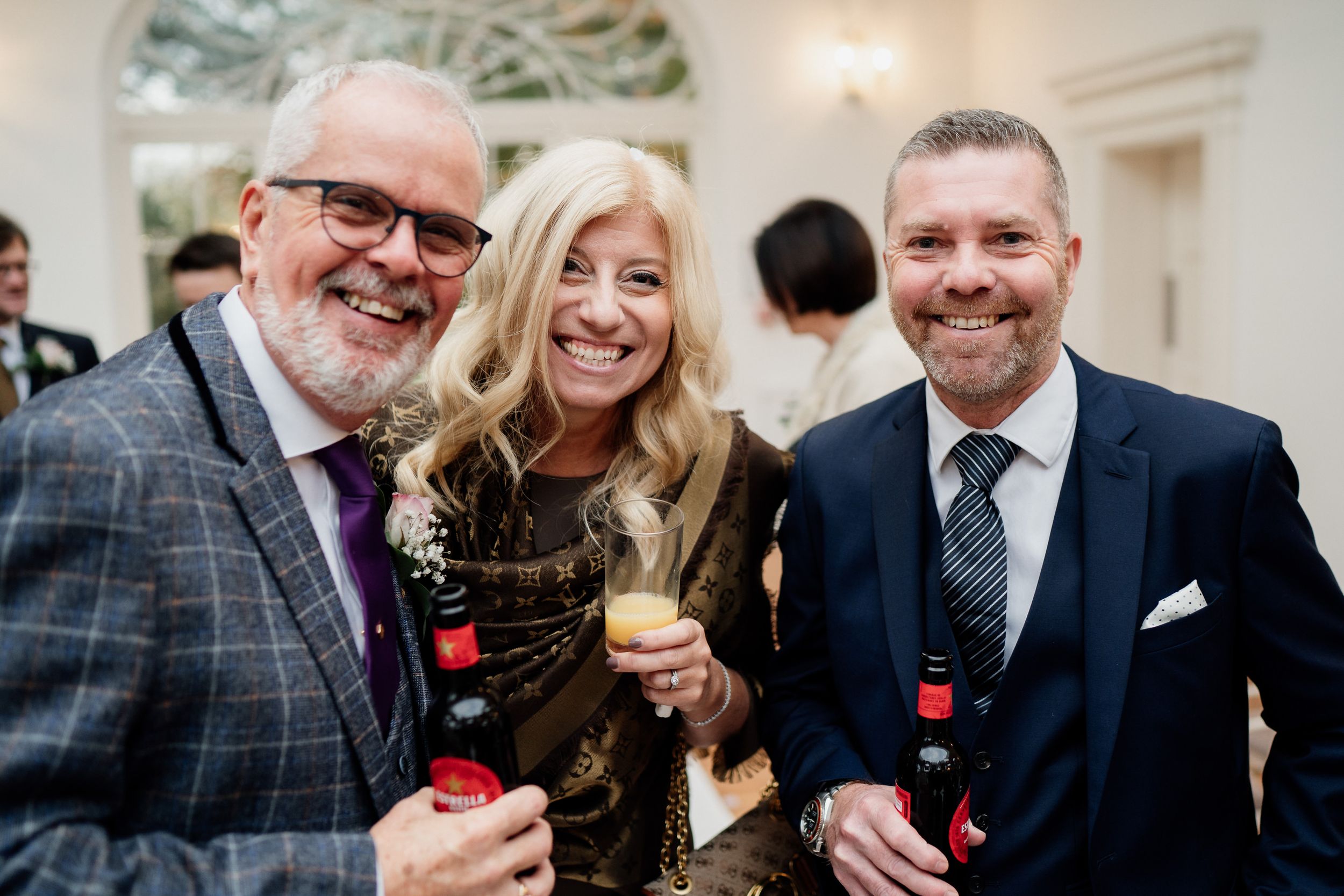 Three wedding guests smile while holding drinks at an elegant indoor reception.