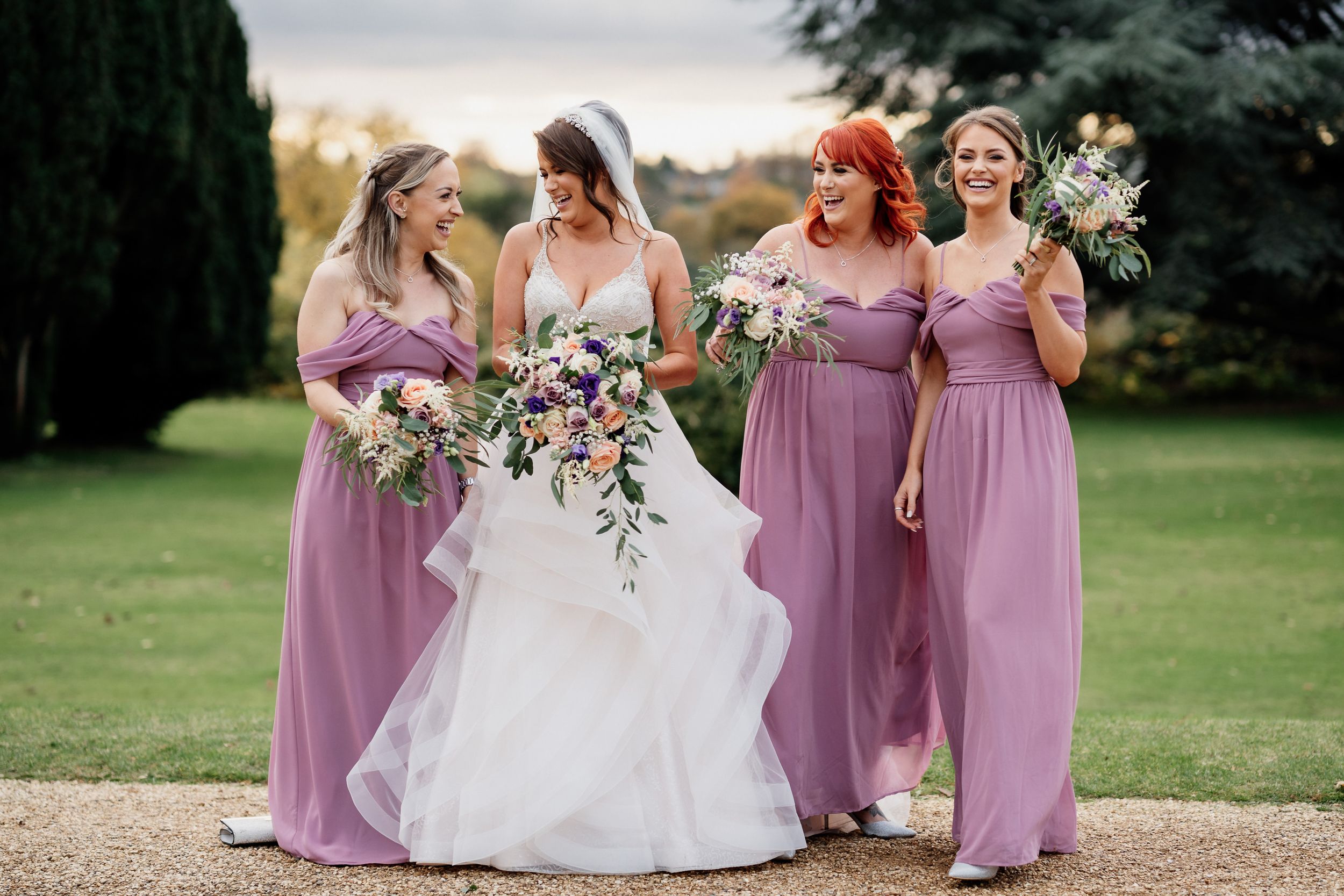 Bride in white gown stands with bridesmaids wearing lavender dresses holding floral bouquets.