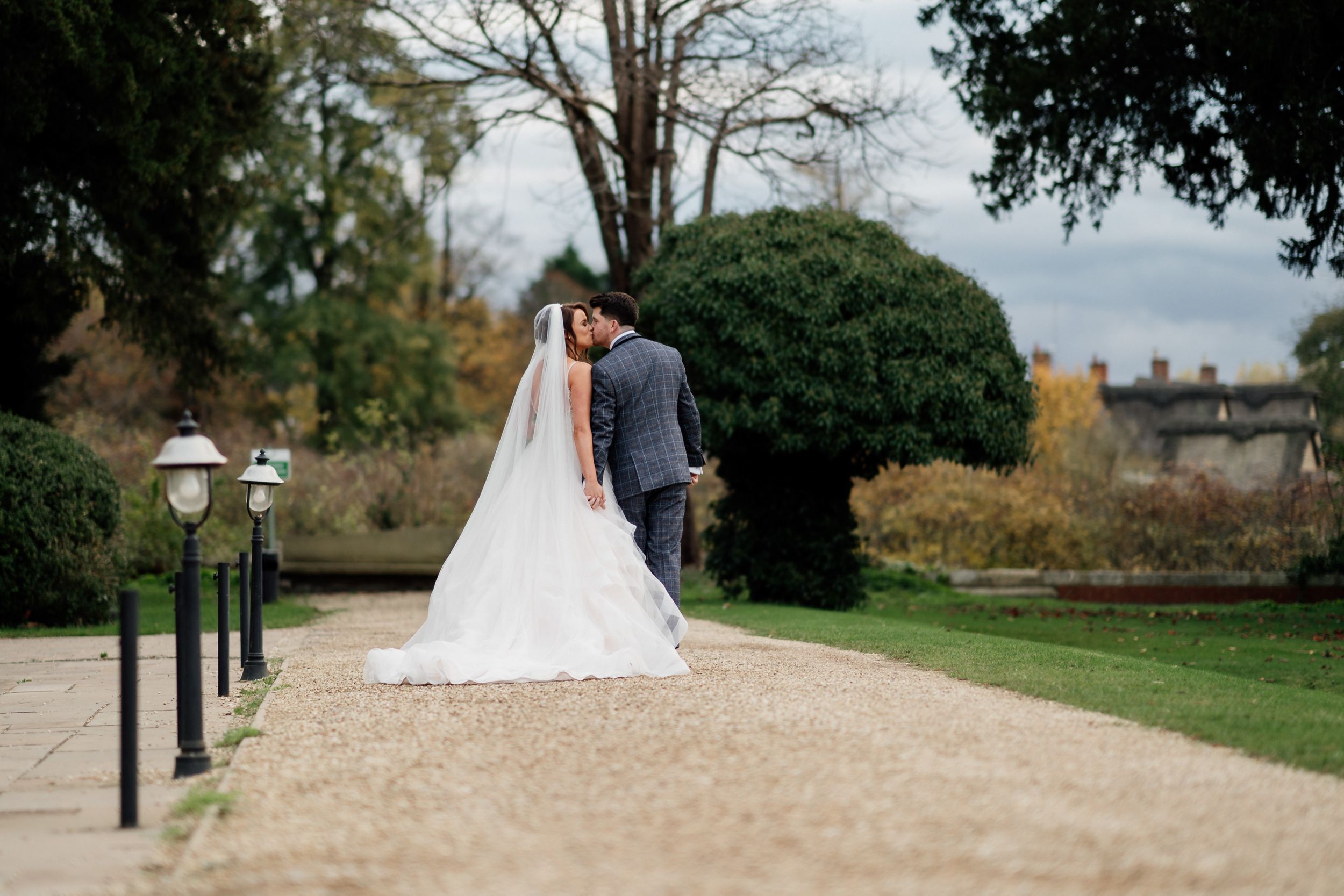 Couple in wedding attire walking along tree-lined garden path.
