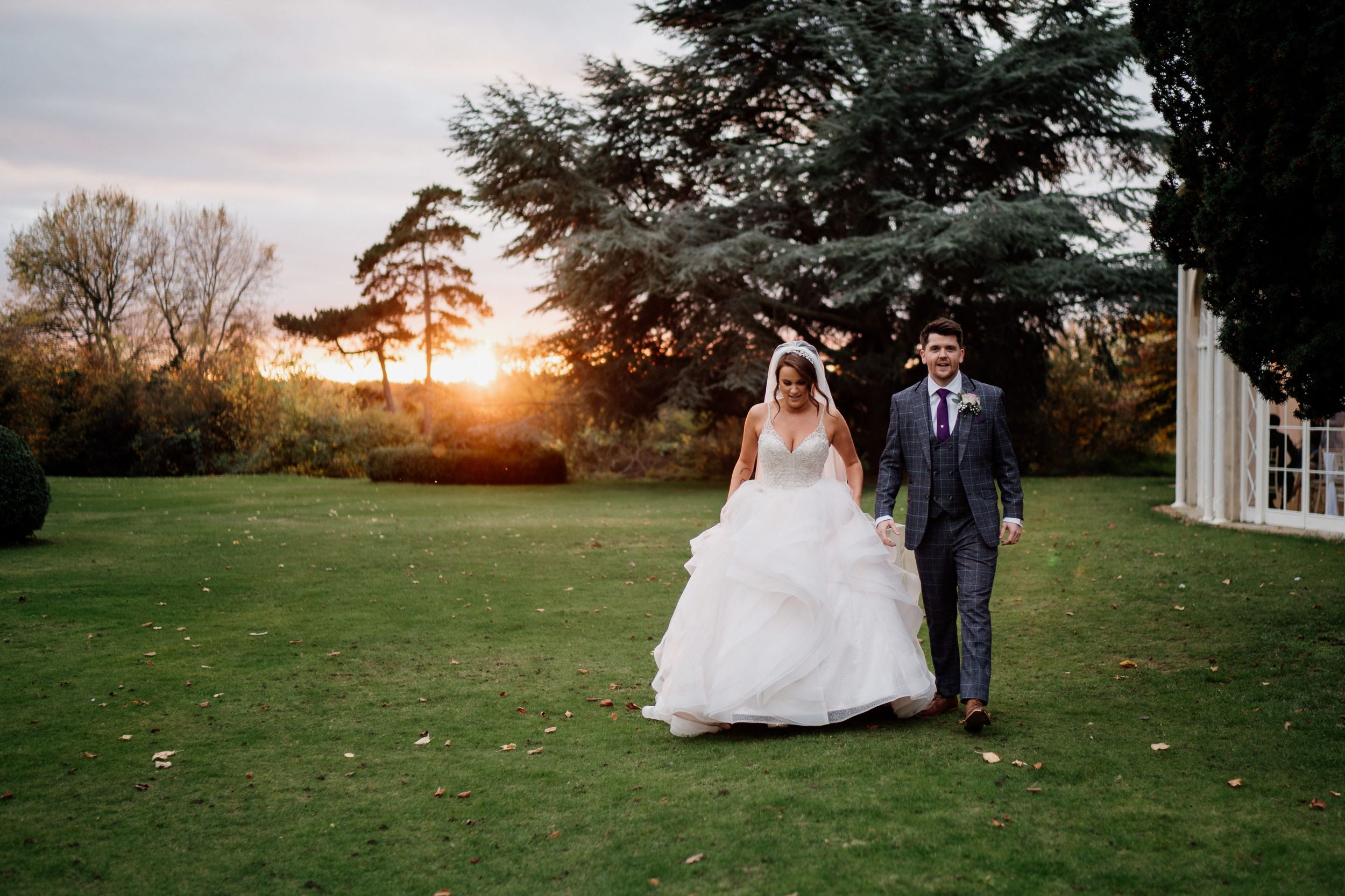 Couple in wedding attire walking on lawn at sunset at Barton Hall with trees in background.