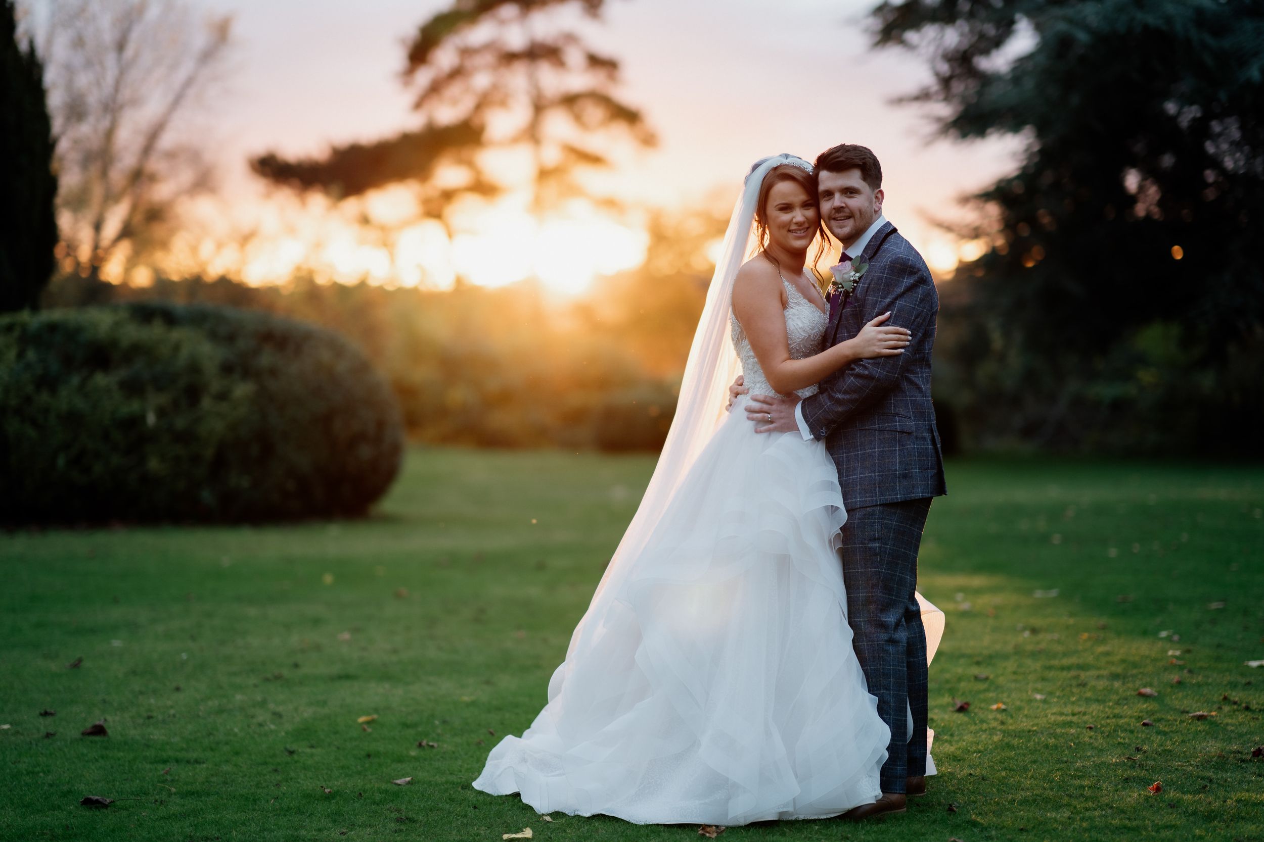 A romantic sunset wedding portrait at Barton Hall shows a couple embracing on a grassy lawn with golden light filtering through trees.