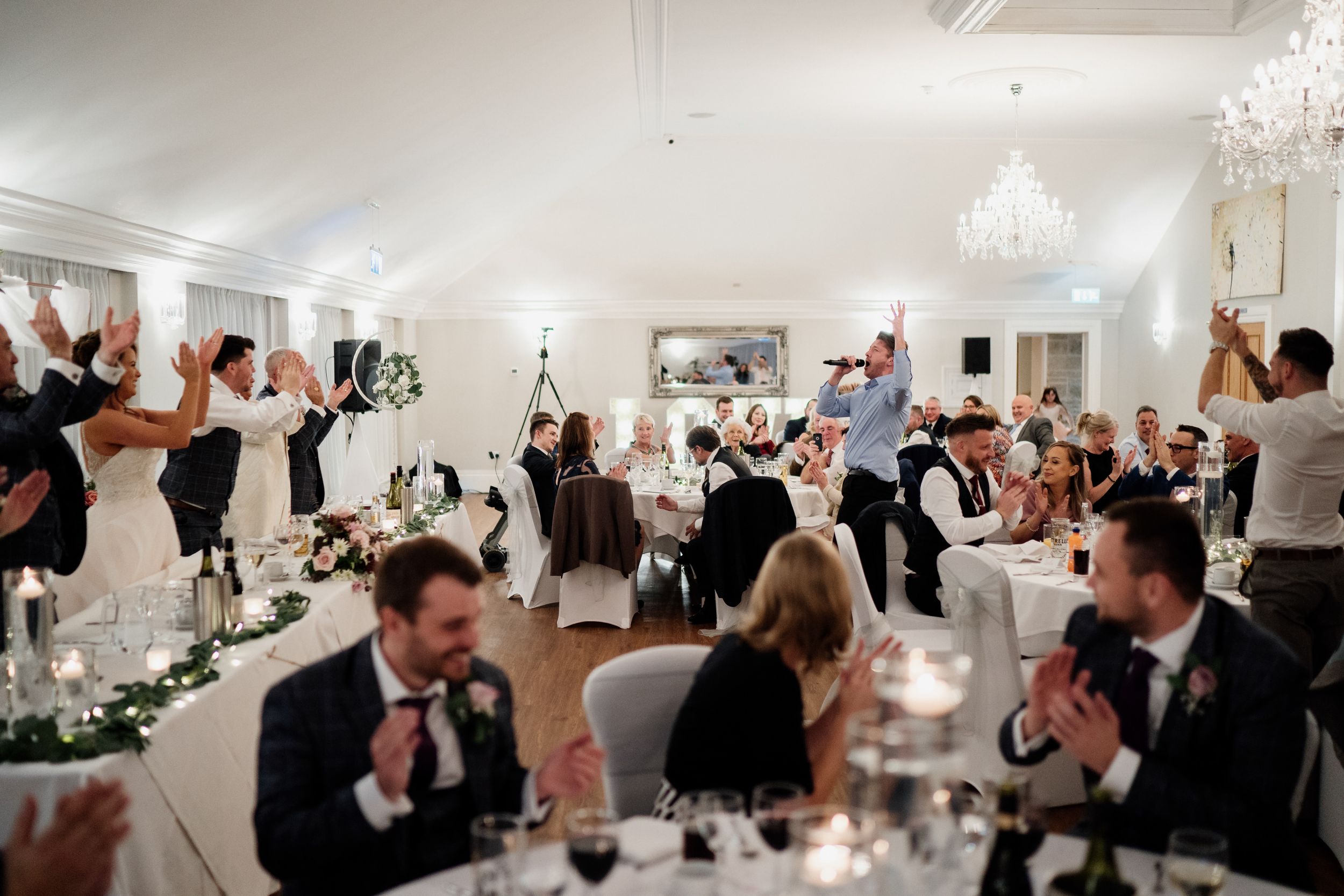 Energetic wedding reception scene with guests cheering and celebrating in a chandelier-lit venue.