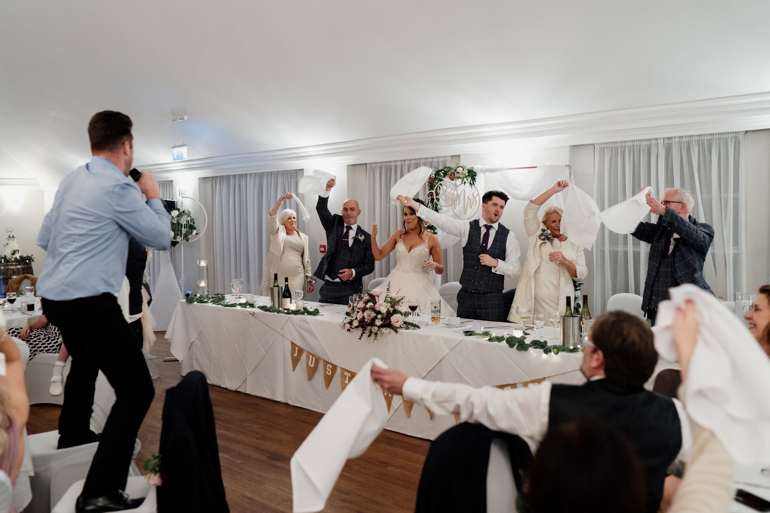 Wedding guests celebrate with enthusiasm during a reception in a white-decorated venue.