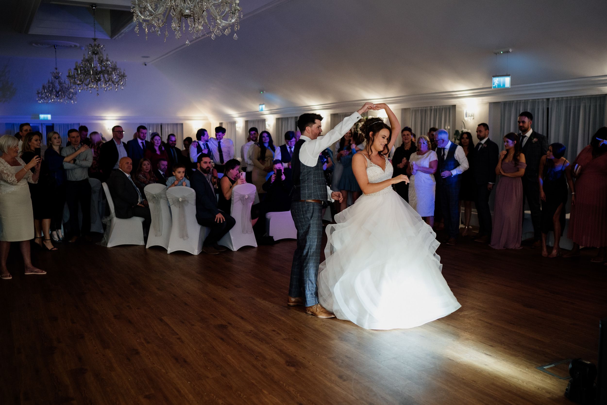 Newlyweds perform their first dance at Barton Hall under spotlights while guests watch in a ballroom setting.