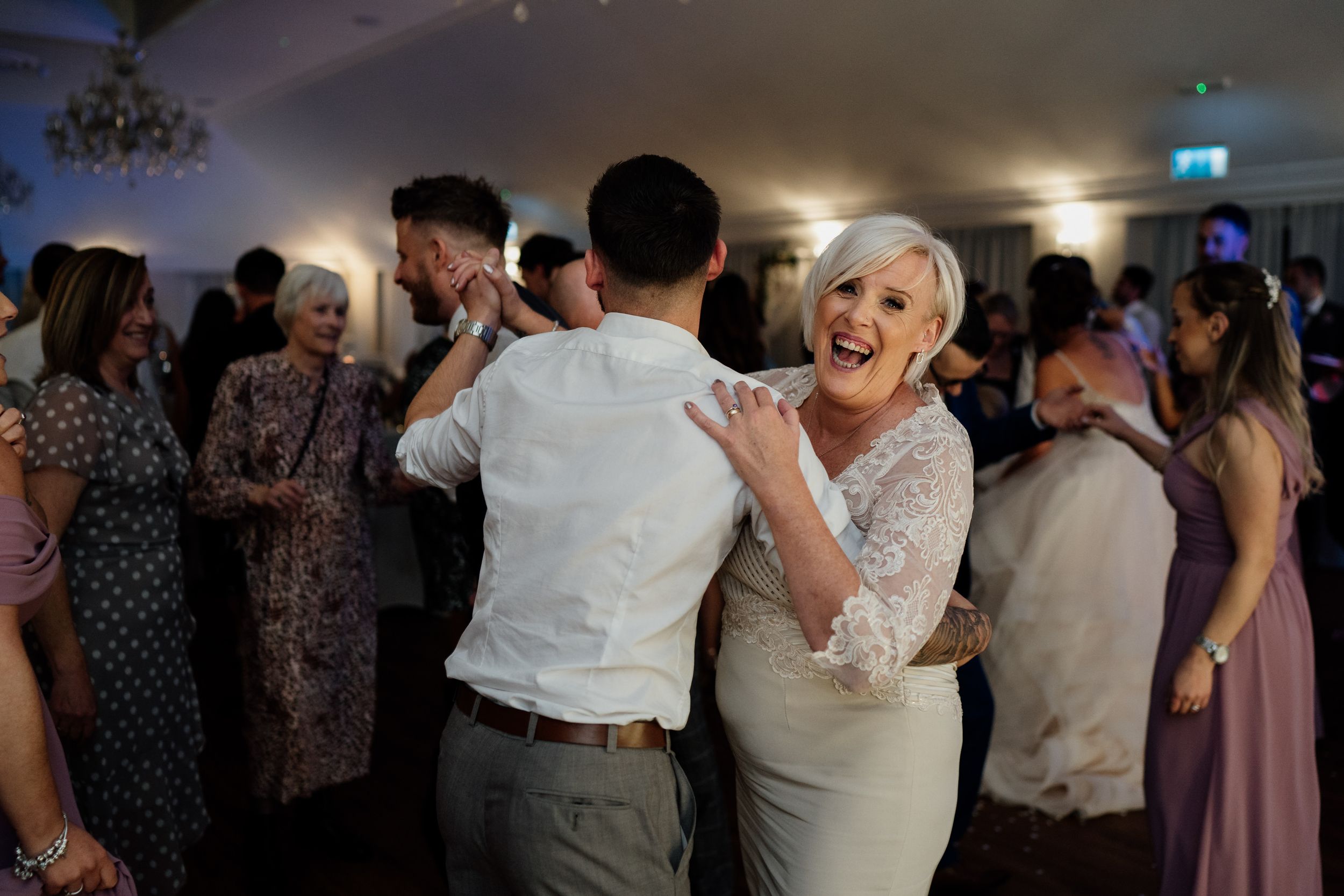 Wedding guests dancing and celebrating at an indoor reception venue with chandelier lighting.