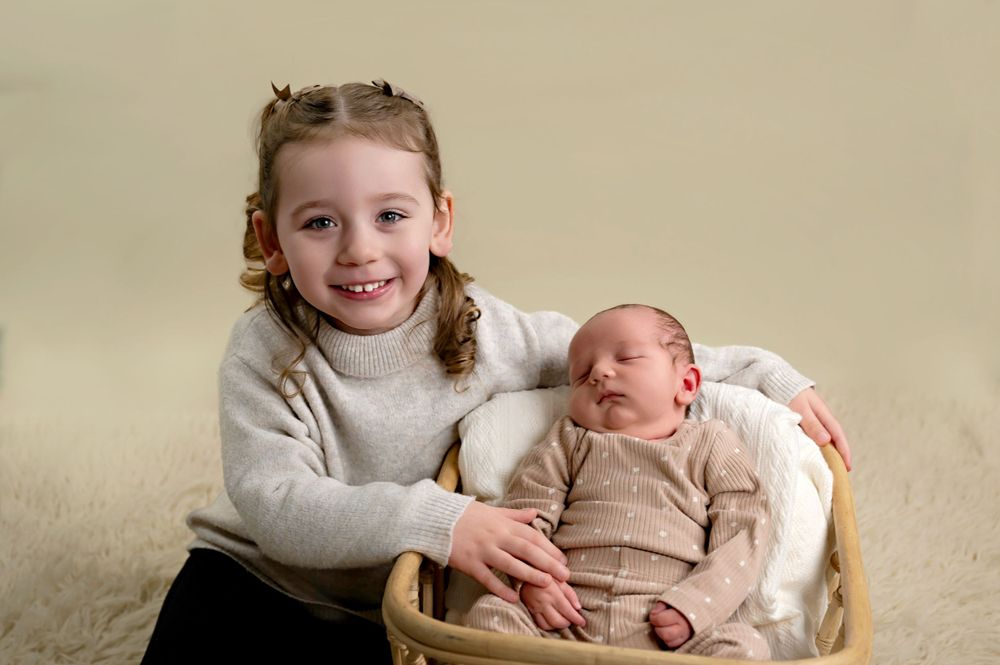 A joyful moment captured as a young child sits with a sleeping newborn baby in a cozy studio setting.