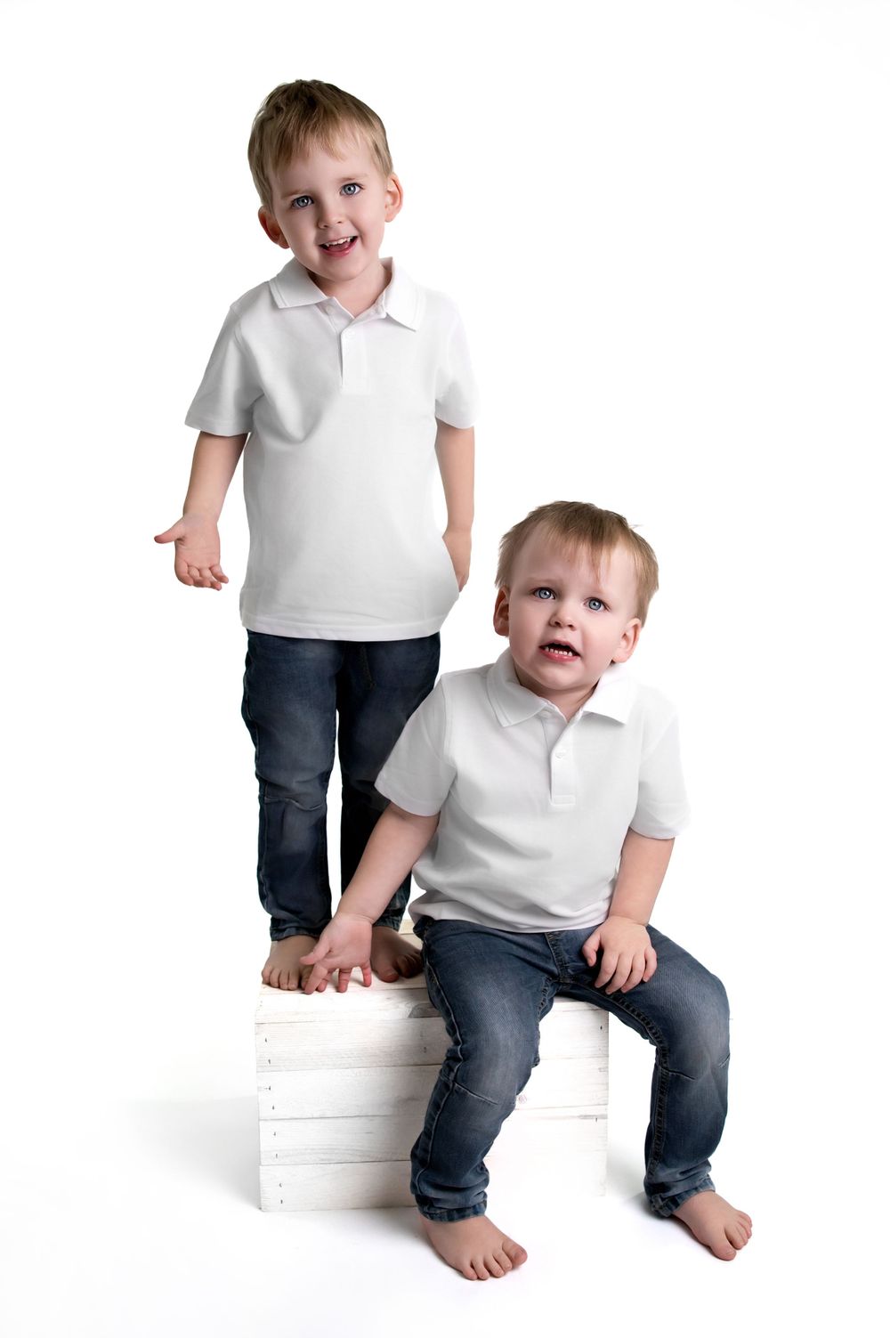 Two young children in white polo shirts and jeans pose casually against a white wooden crate backdrop.