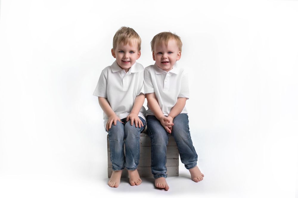 Two small children in matching white shirts and jeans sit together laughing against a white backdrop.