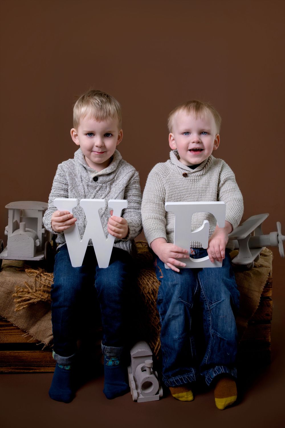 Two children in sweaters sitting side by side holding white decorative letters on a brown sofa.