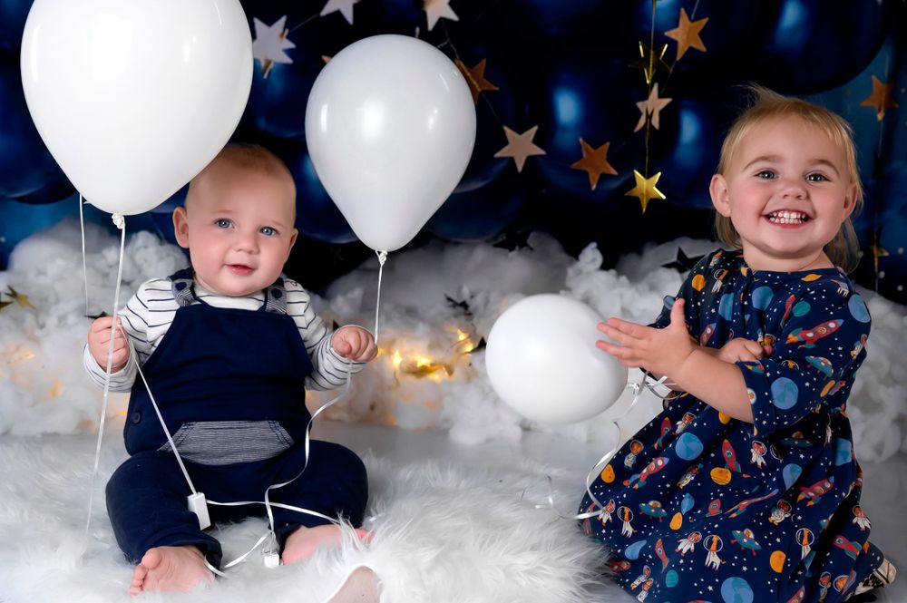Two children pose with white balloons against a dark backdrop with stars and cloud decorations in a studio setting.