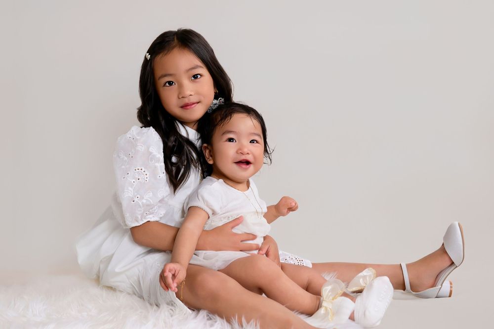 Two people in white outfits sit together on a white backdrop for a portrait session.