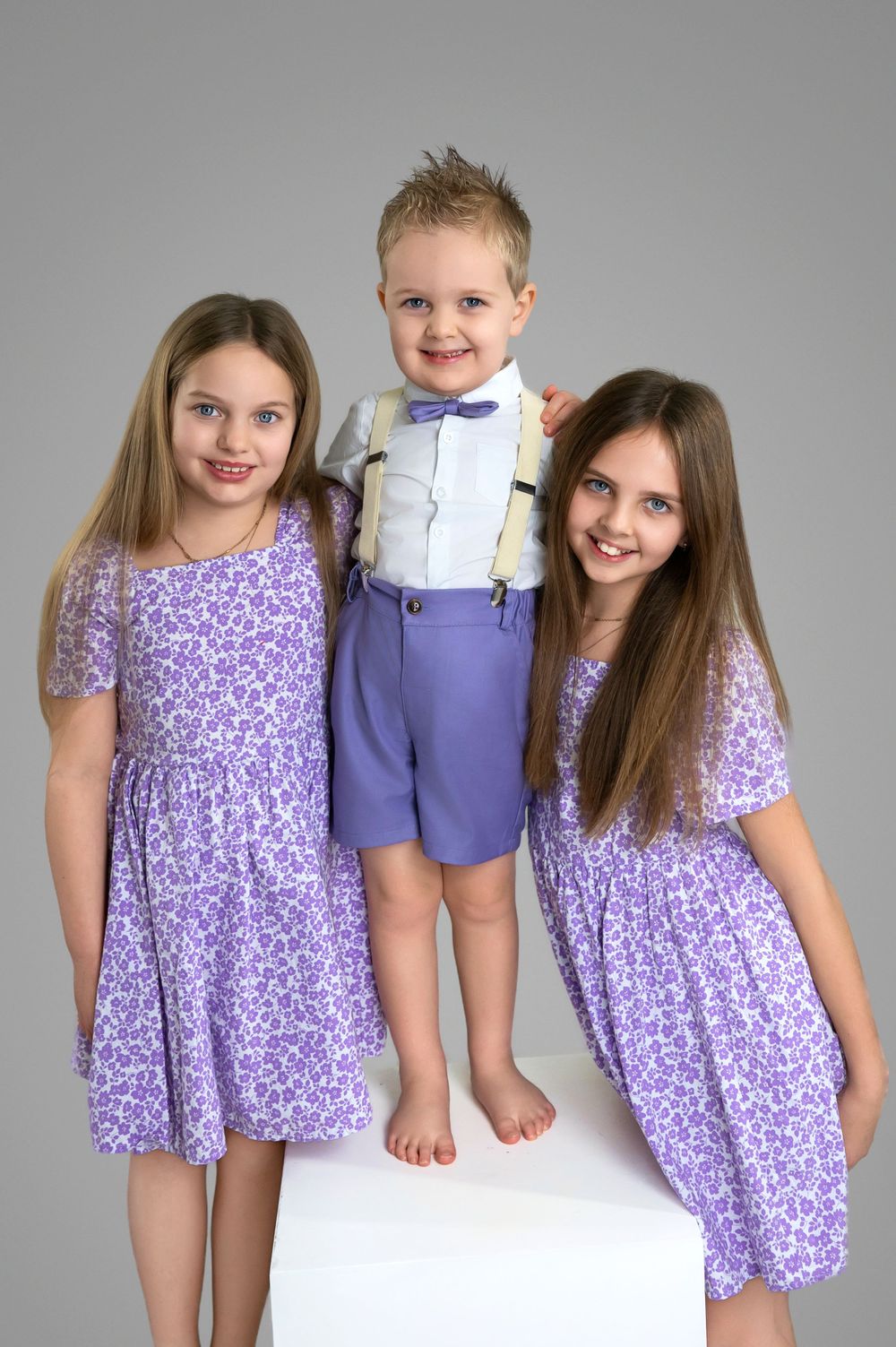 Three people in coordinating purple floral dresses and blue outfit pose together on white backdrop for formal portrait.